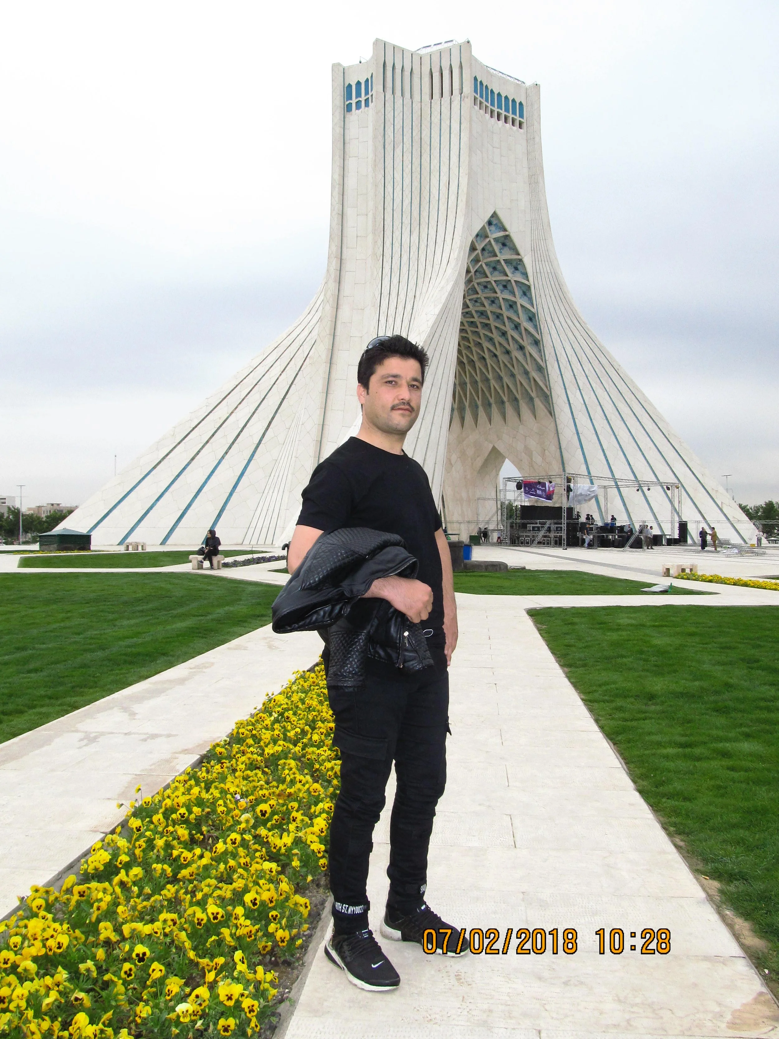 A man standing on a sidewalk in front of the Azadi Tower in Tehran, Iran, holding a black jacket. The tower is a large, white, modern architectural monument with a curved structure. The sky is overcast, and there are yellow flowers along the sidewalk.