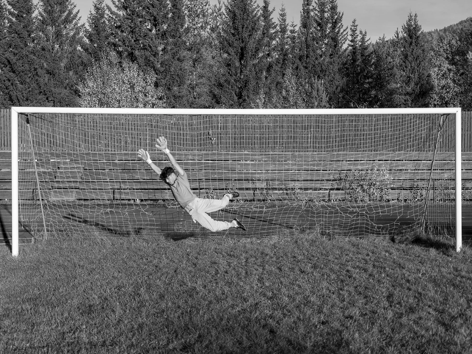 A young boy in mid-air diving or falling near a soccer goal on a grassy field, with a background of trees and a wooden fence.