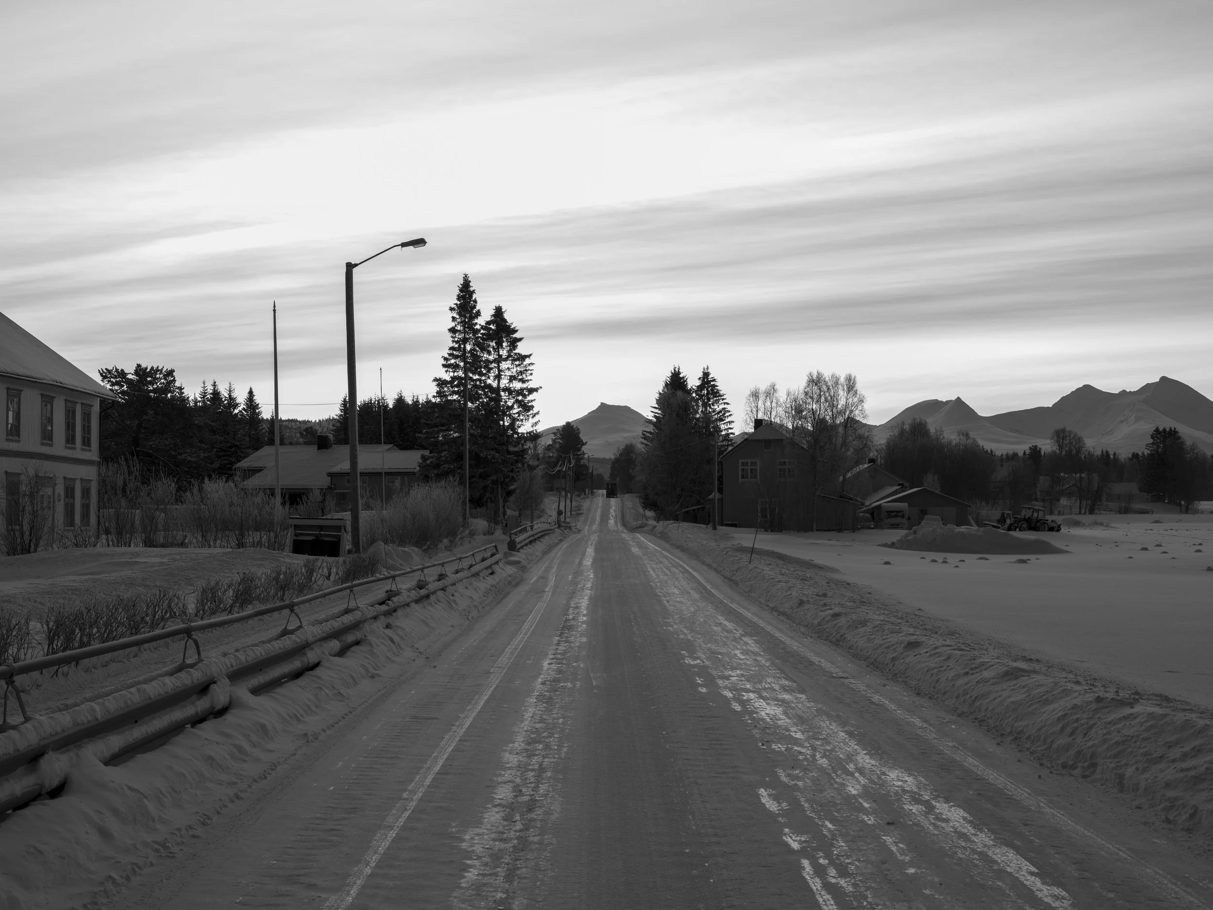 Black and white photo of a snow-covered rural street with houses, trees, streetlights, and mountains in the background.
