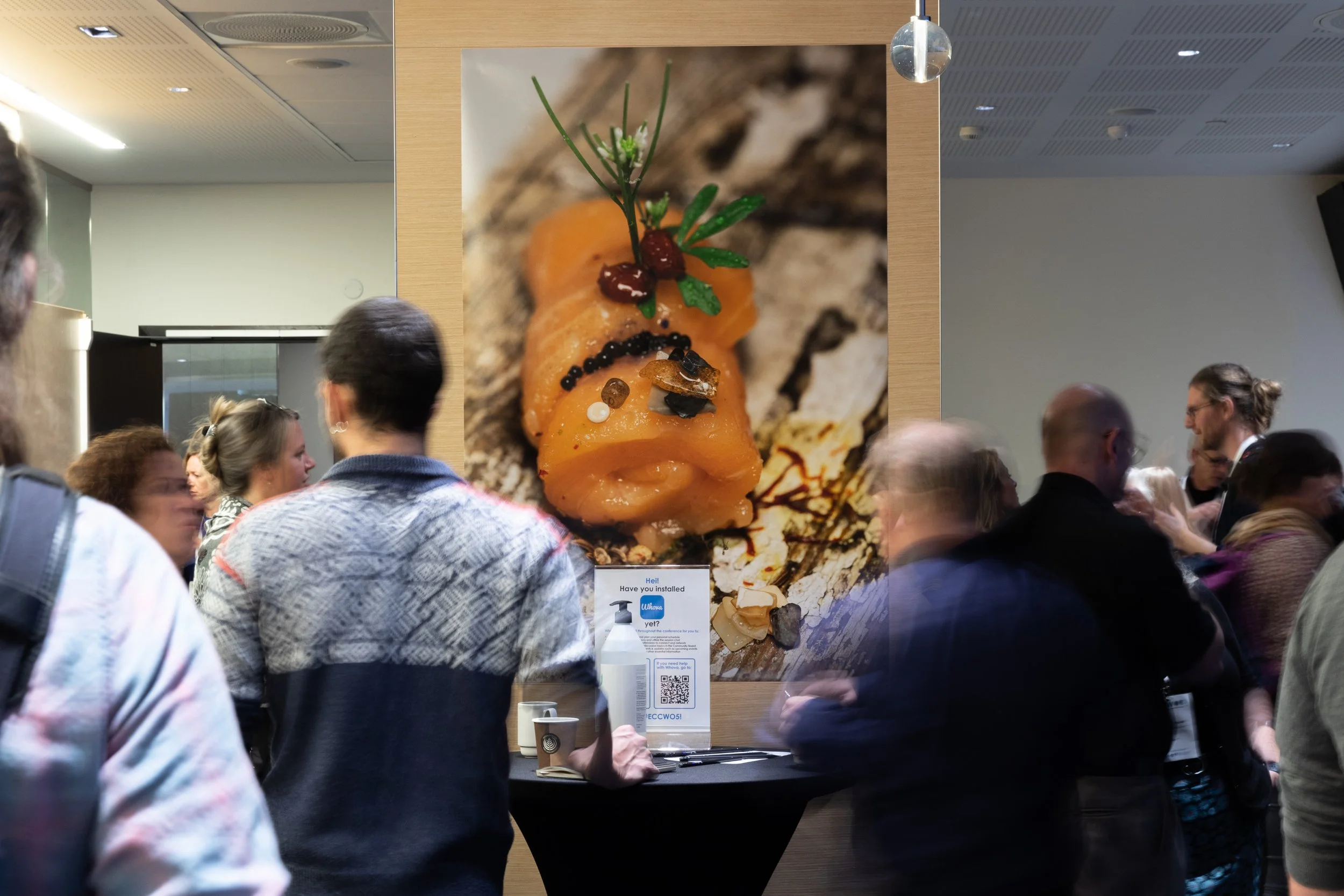 Large display of gourmet dish with orange-colored fish or seafood, garnished with herbs, spices, and sauces, surrounded by a crowd of people at an event or exhibition.