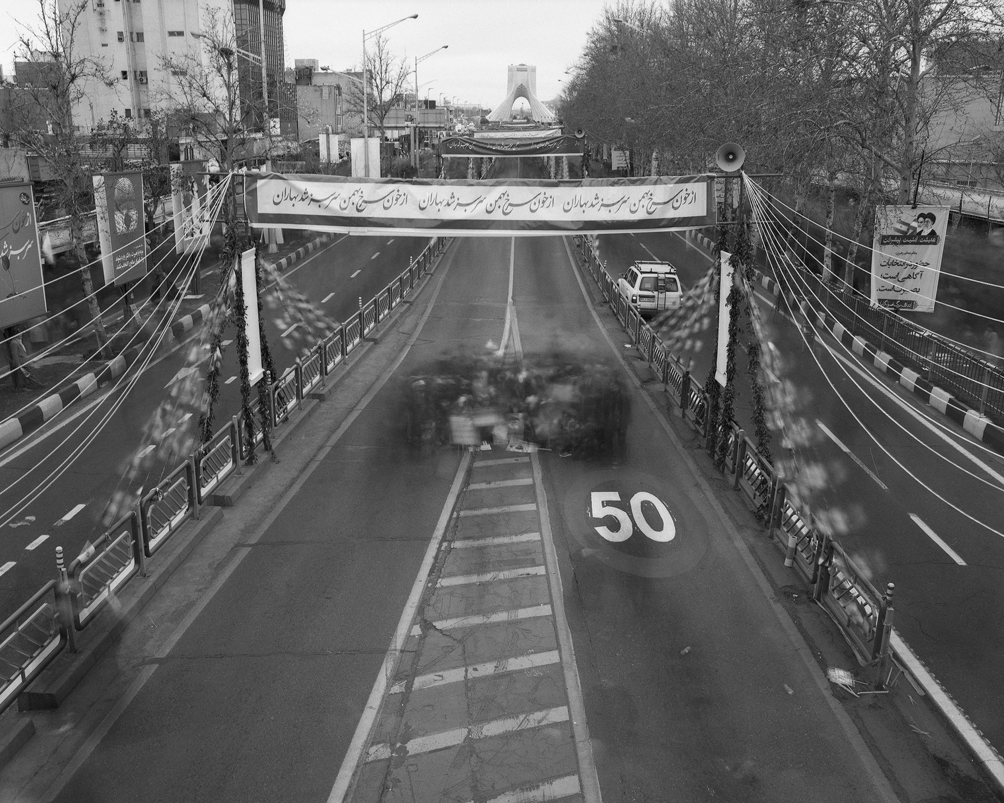 View of a street with a speed limit sign of 50, parked cars, and a bridge in the distance, seen from behind a glass window with reflections.