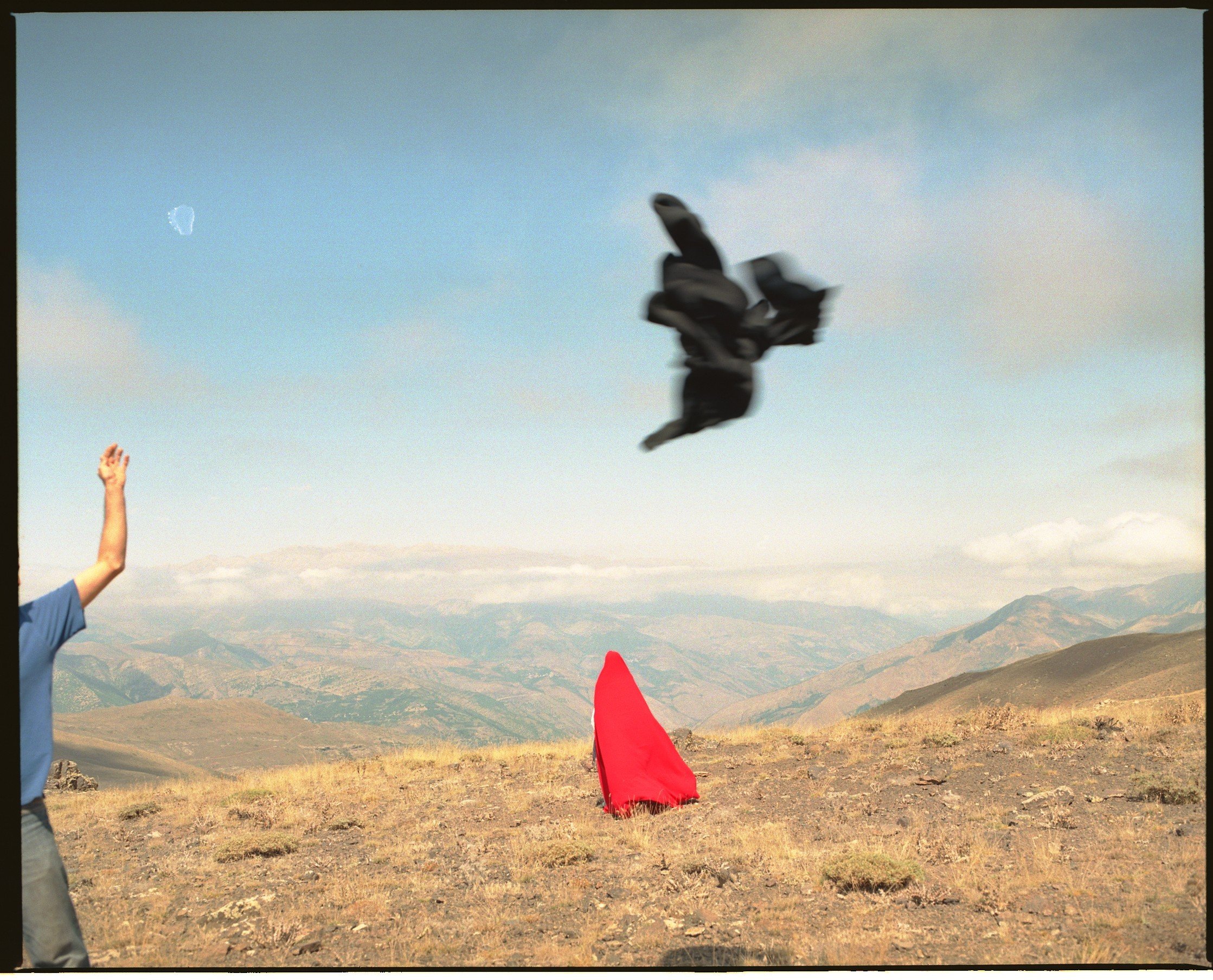 A person throwing a black jacket or piece of clothing into the air in an open landscape with mountains in the background, a red object on the ground, and a bird flying in the sky.