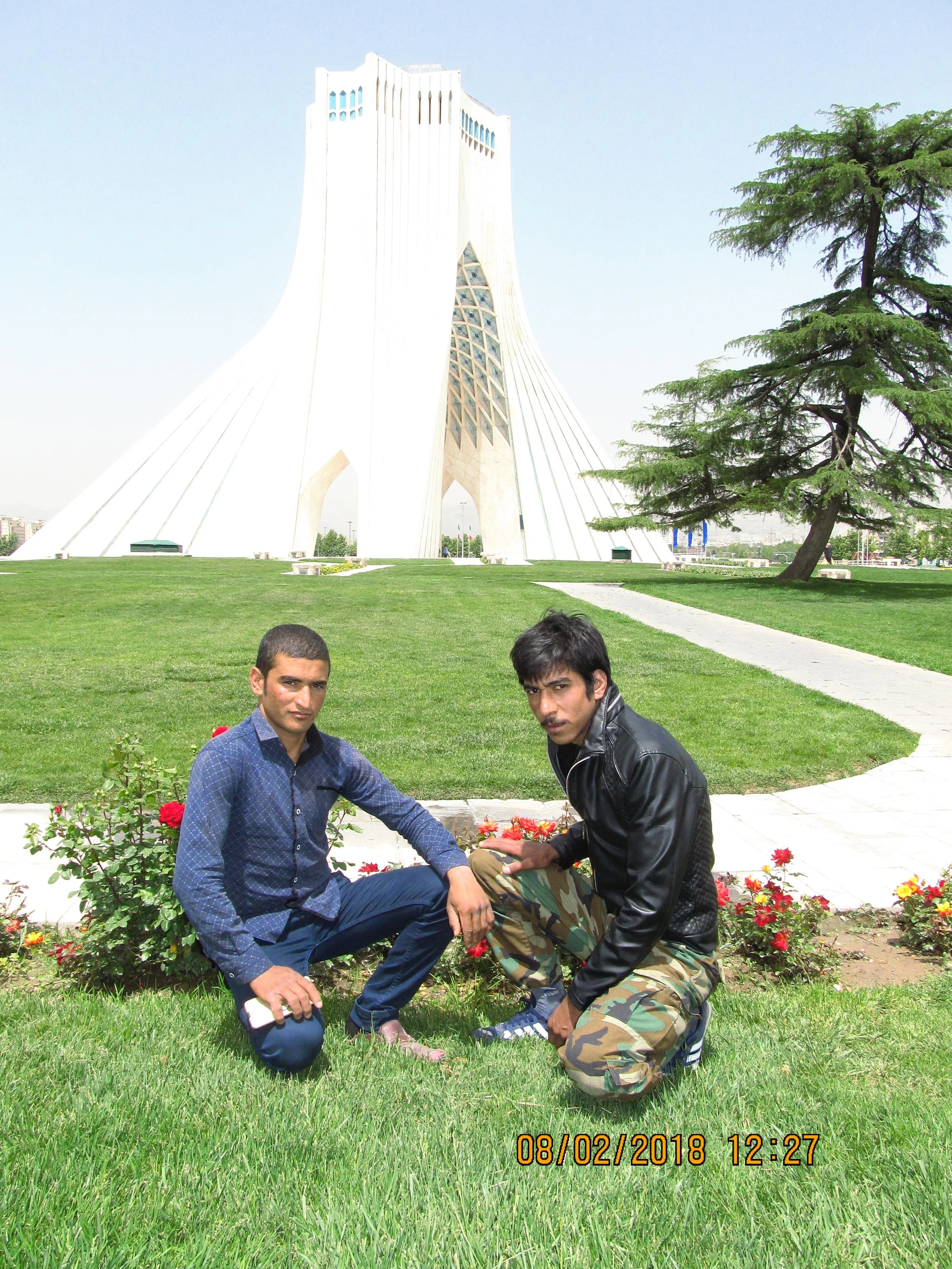 Two young men kneeling on the grass in front of a flower bed with a monument and a tree in the background.