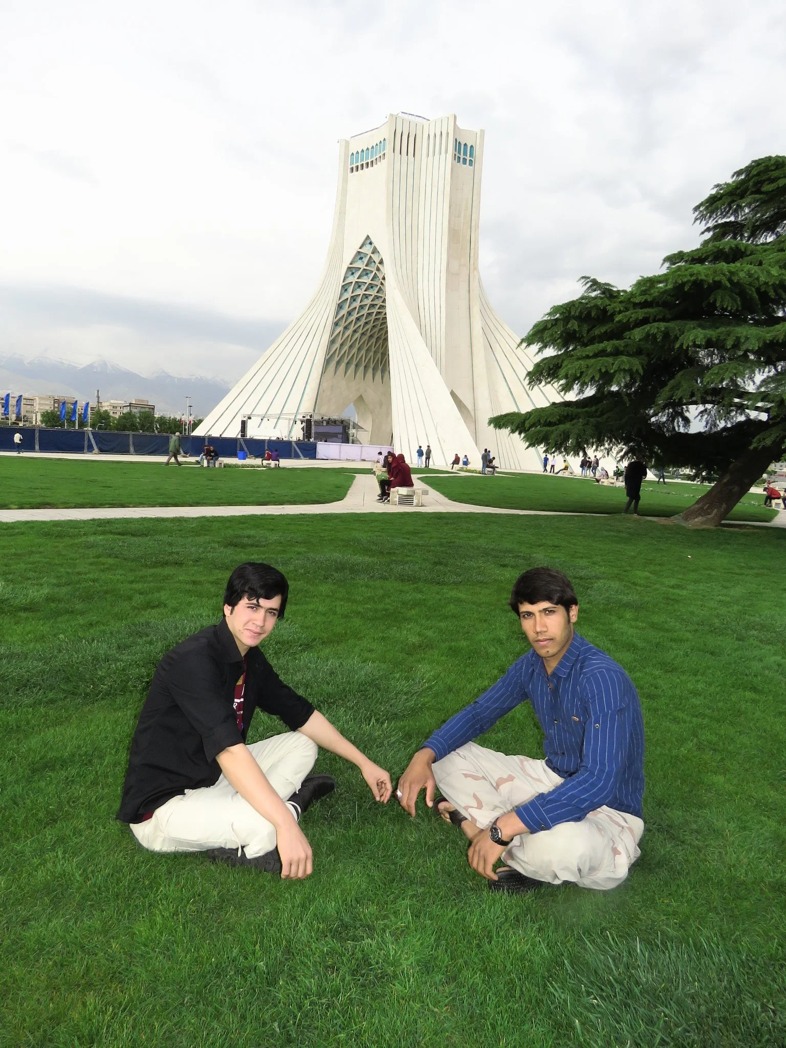 Two young men sitting on green grass in front of the Azadi Tower in Tehran, Iran, with the tower’s distinctive white structure and cloudy sky in the background.