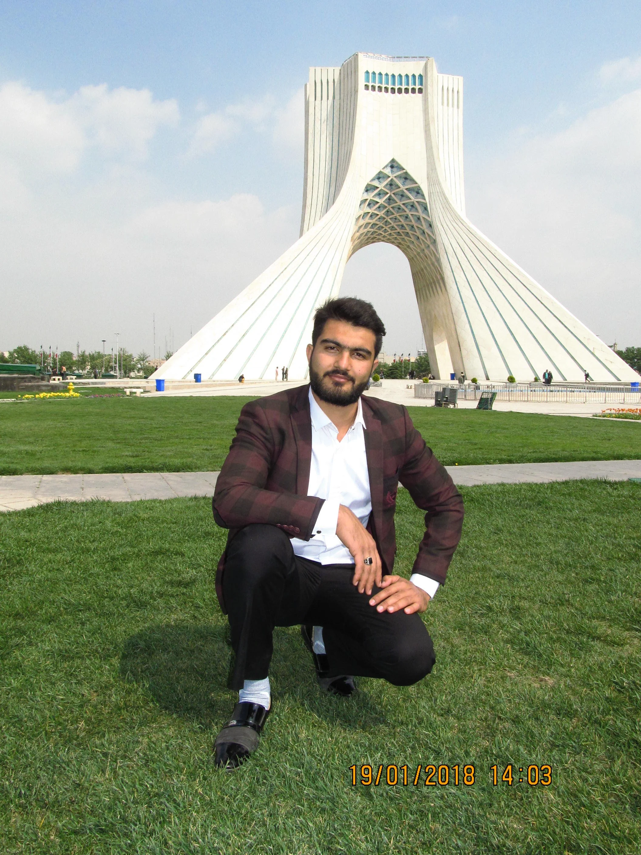 A man in a suit crouching on grass in front of a large white monument with unique architectural design, under a cloudy sky.