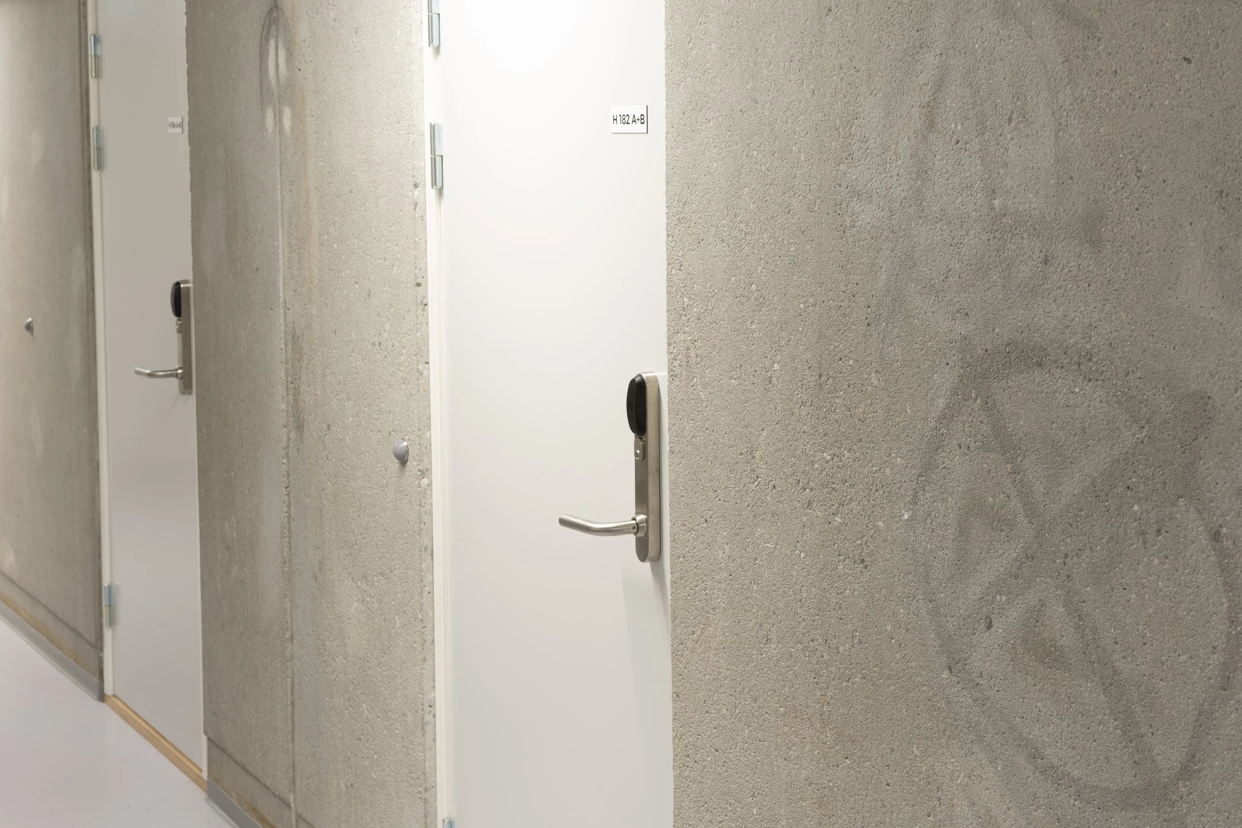 A corridor with three closed doors on textured concrete walls, each door has a handle and a lock, with a sign on the middle door reading 'H 182 A+B'.