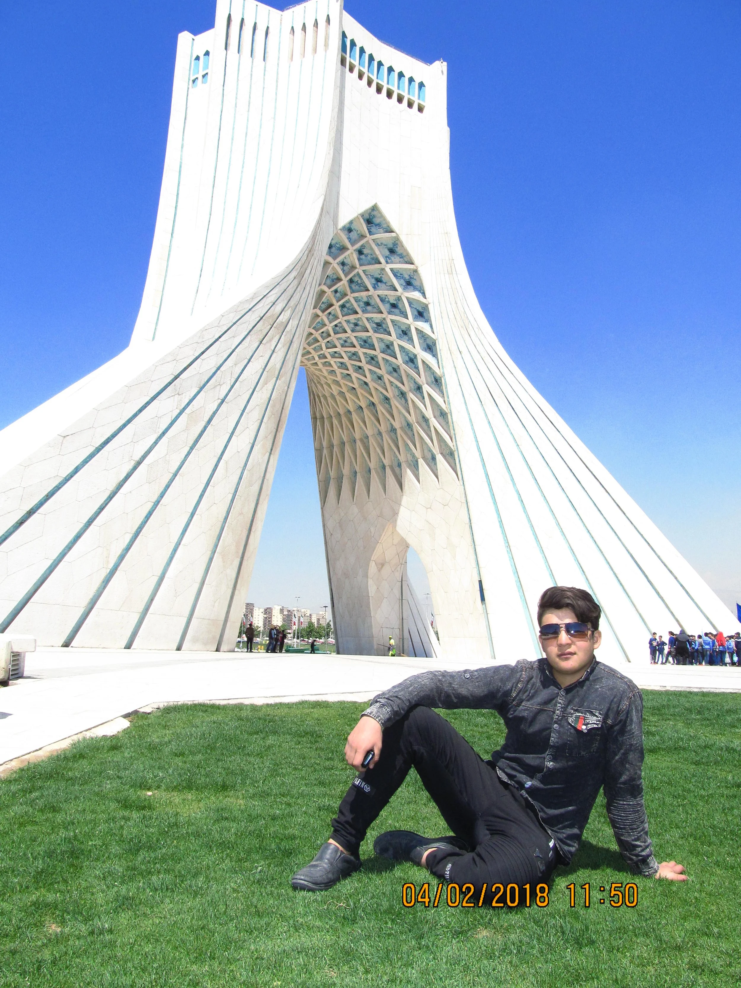 Young man wearing sunglasses and black clothing sitting on grass in front of Azadi Tower in Tehran, Iran, on a sunny day.