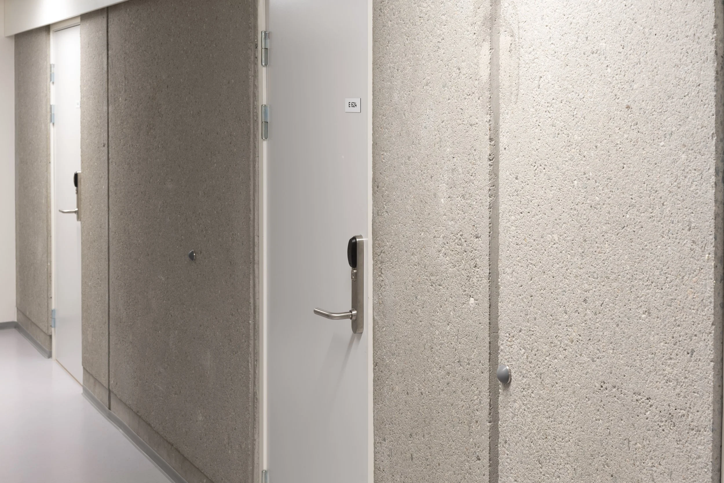 A corridor with textured beige walls and several white doors, one of which has a black handle and a small label marked 'E 624'.