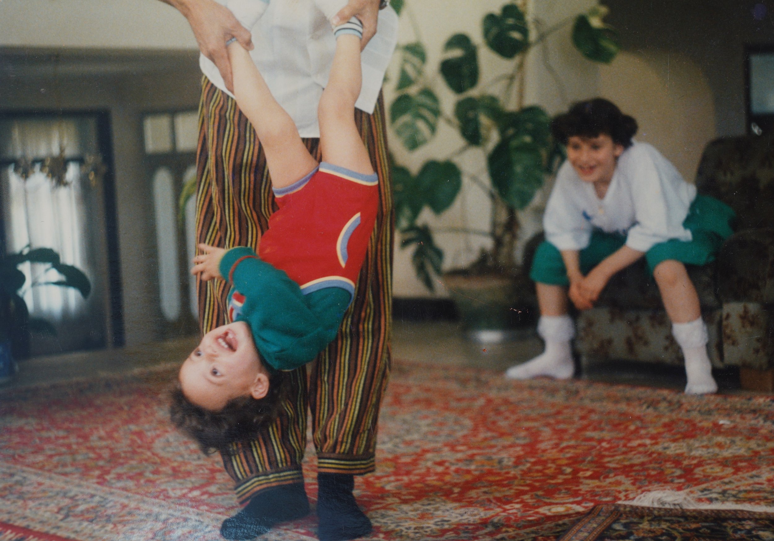 A young boy hanging upside down from an adult's hands, laughing, while a girl sits on a couch in the background smiling. They are indoors with a patterned carpet and large houseplants.
