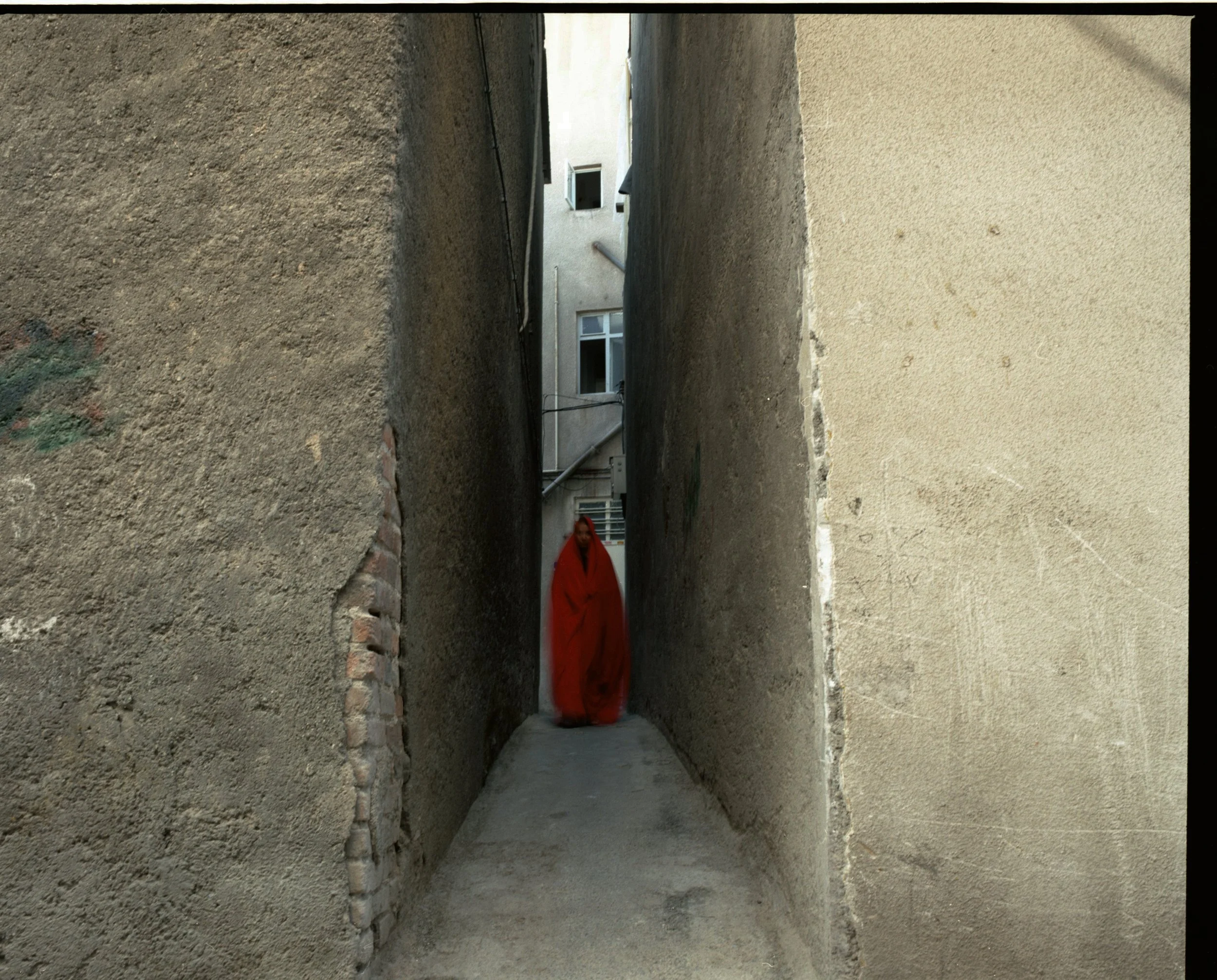 A person in a red cloak walking through a narrow alley between two tall, textured walls in an urban setting.