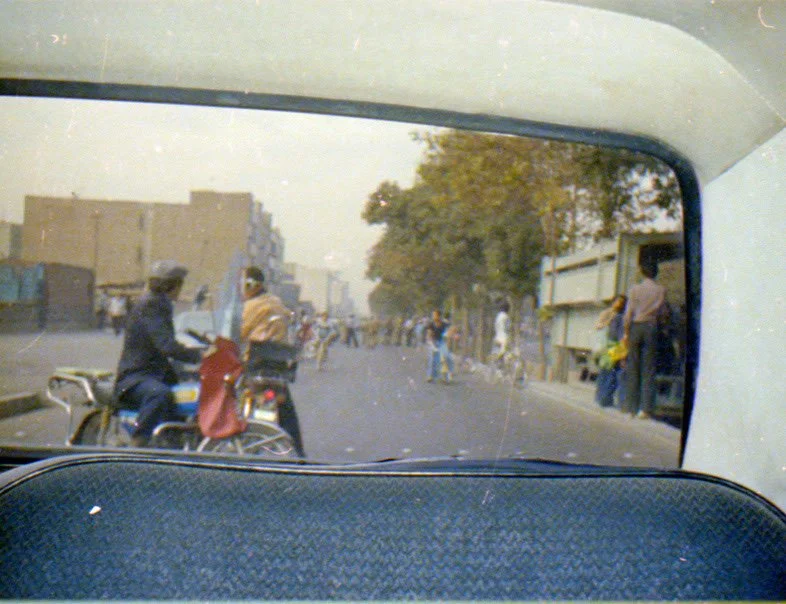 View of a street scene through a car window, showing motorcycles and pedestrians on the street, with buildings and trees in the background.