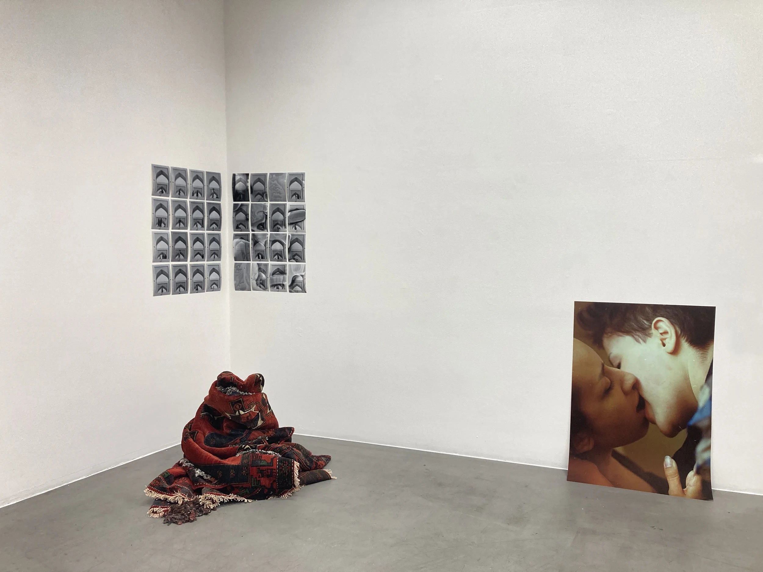 An art installation featuring a pile of rolled-up and folded red and black patterned fabric on the floor in a corner of a room with white walls. On the wall, two sets of small black-and-white photo collages are displayed, and a large color photograph of two people kissing is propped against the right wall.