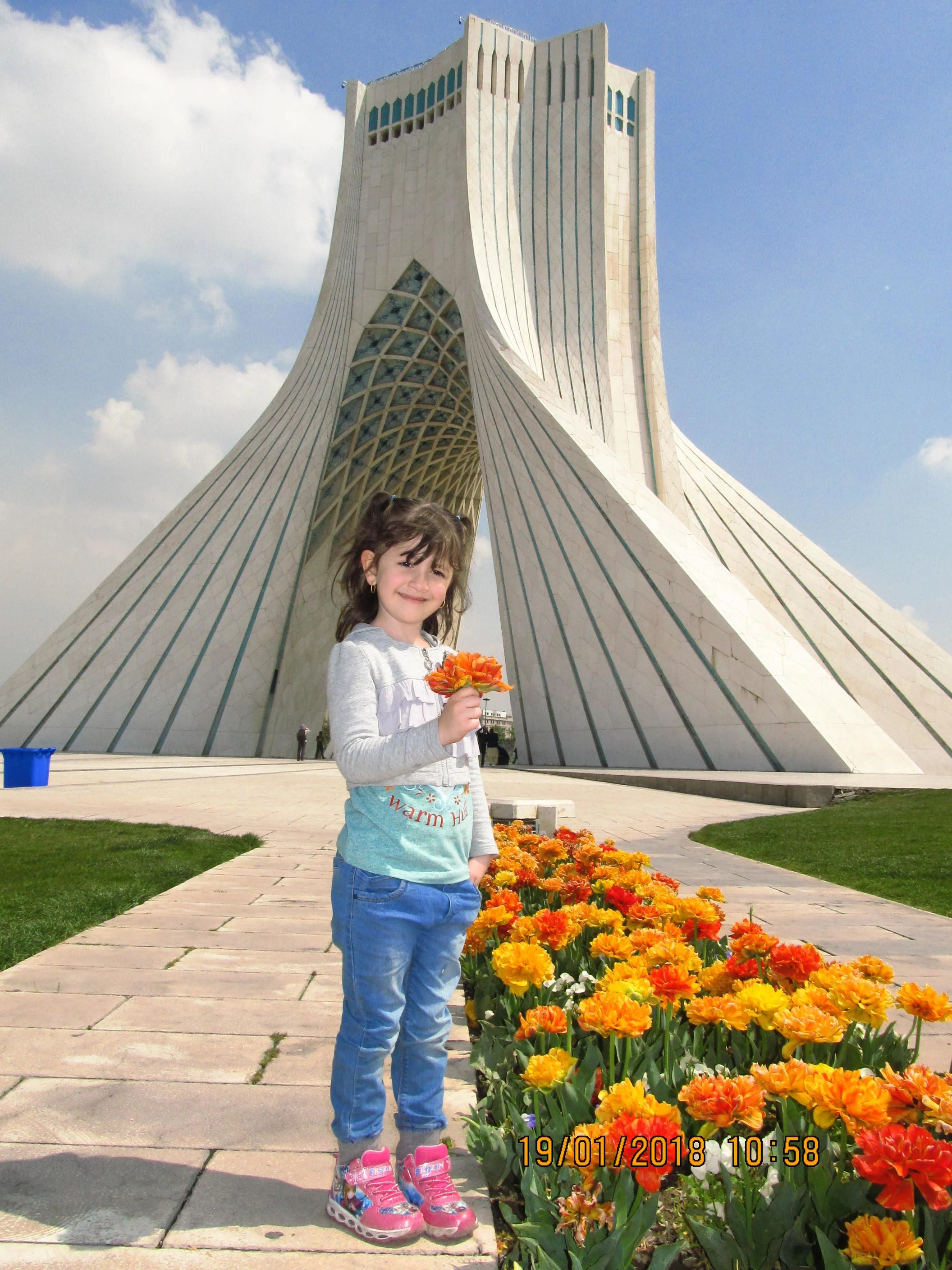 Young girl in gray sweatshirt and blue jeans holding a flower, standing near a bed of orange and yellow flowers in front of a modern white building with unique architectural design on a partly cloudy day.