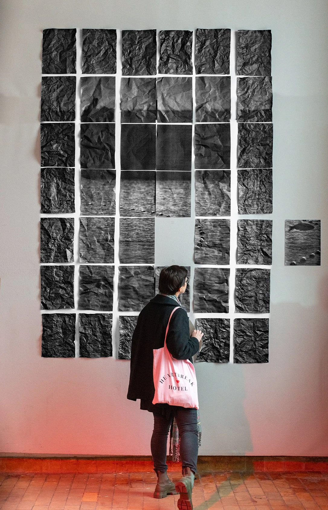A person with short hair, wearing glasses, a black coat, and gray boots, stands in front of a wall displaying an art piece composed of crumpled black and white paper arranged in a grid. The art features black and white photographs of water and rocks, with some smaller photographs on the right side.