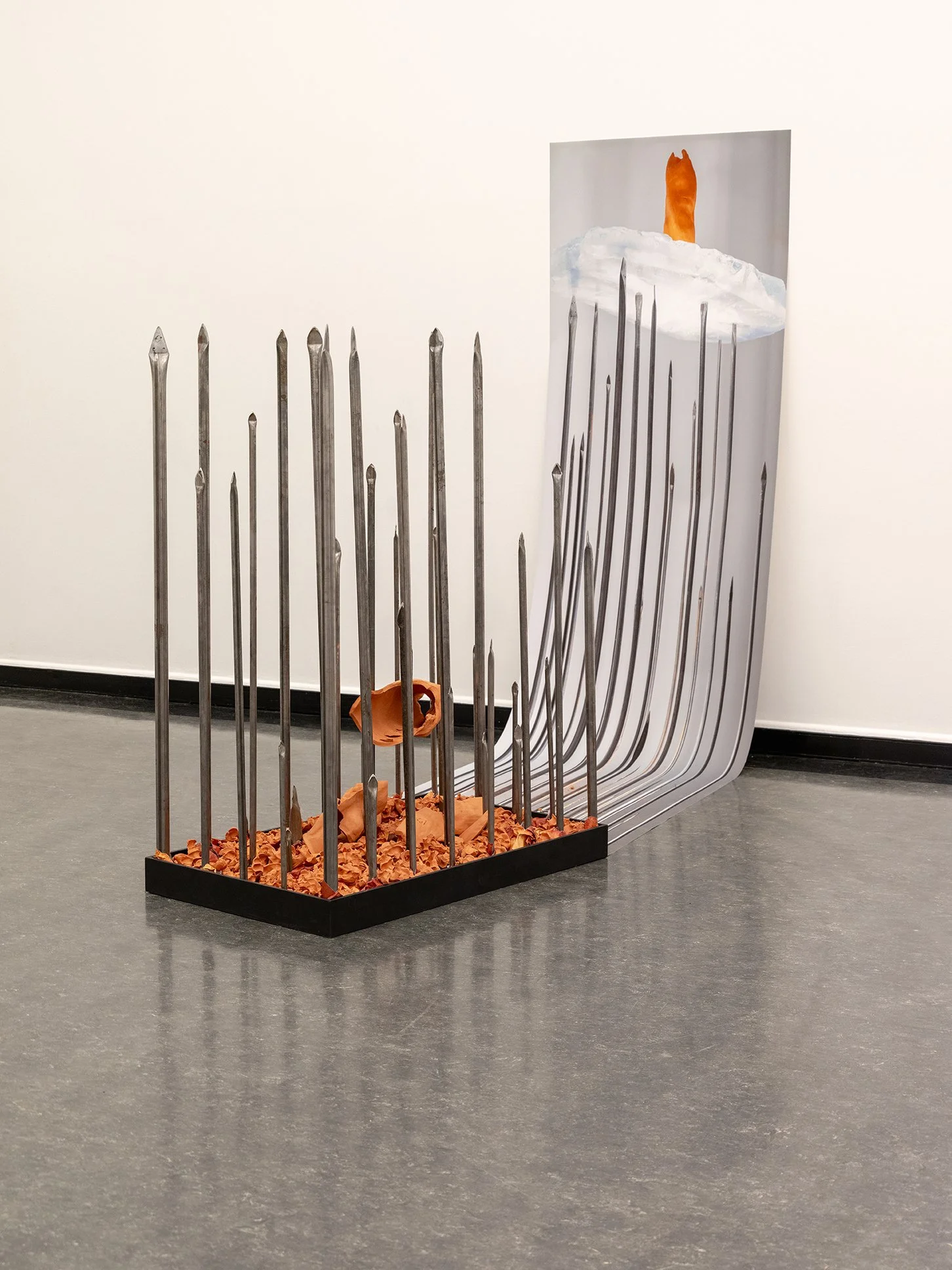 An art installation featuring a collection of large metal nails arranged vertically on a black base with orange crumpled paper, leading into a painted depiction of a candle on a melting snow hillside.