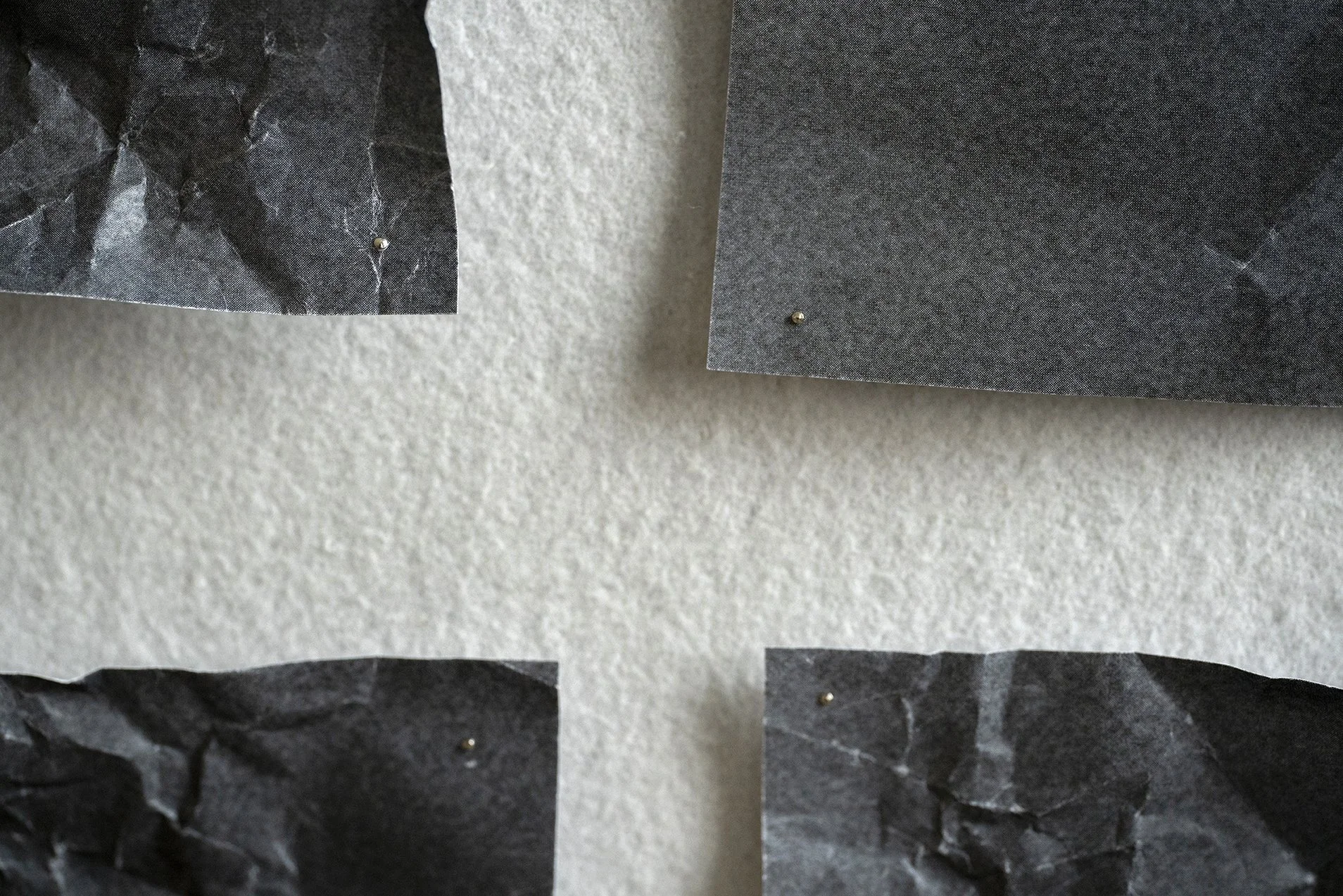 Close-up of four sheets of black and gray textured paper pinned to a white wall, some crumpled, with small metal pins.