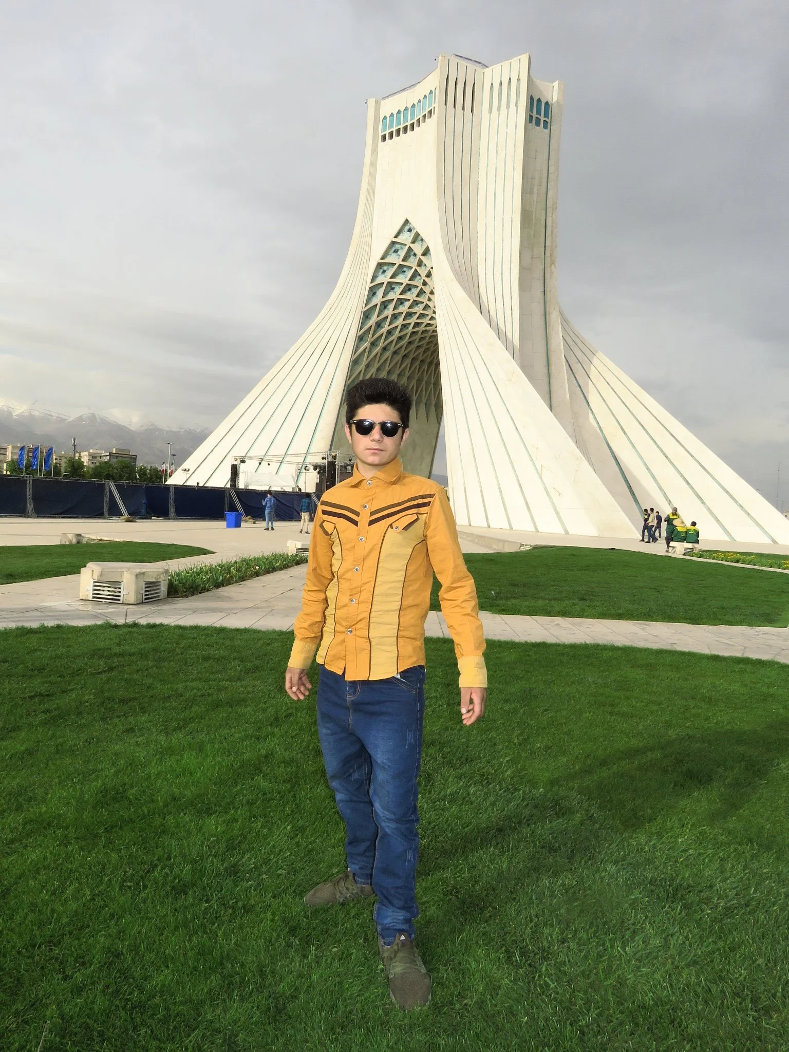 A young man wearing sunglasses, a yellow shirt, and jeans standing on a green lawn in front of the Azadi Tower in Tehran, Iran.