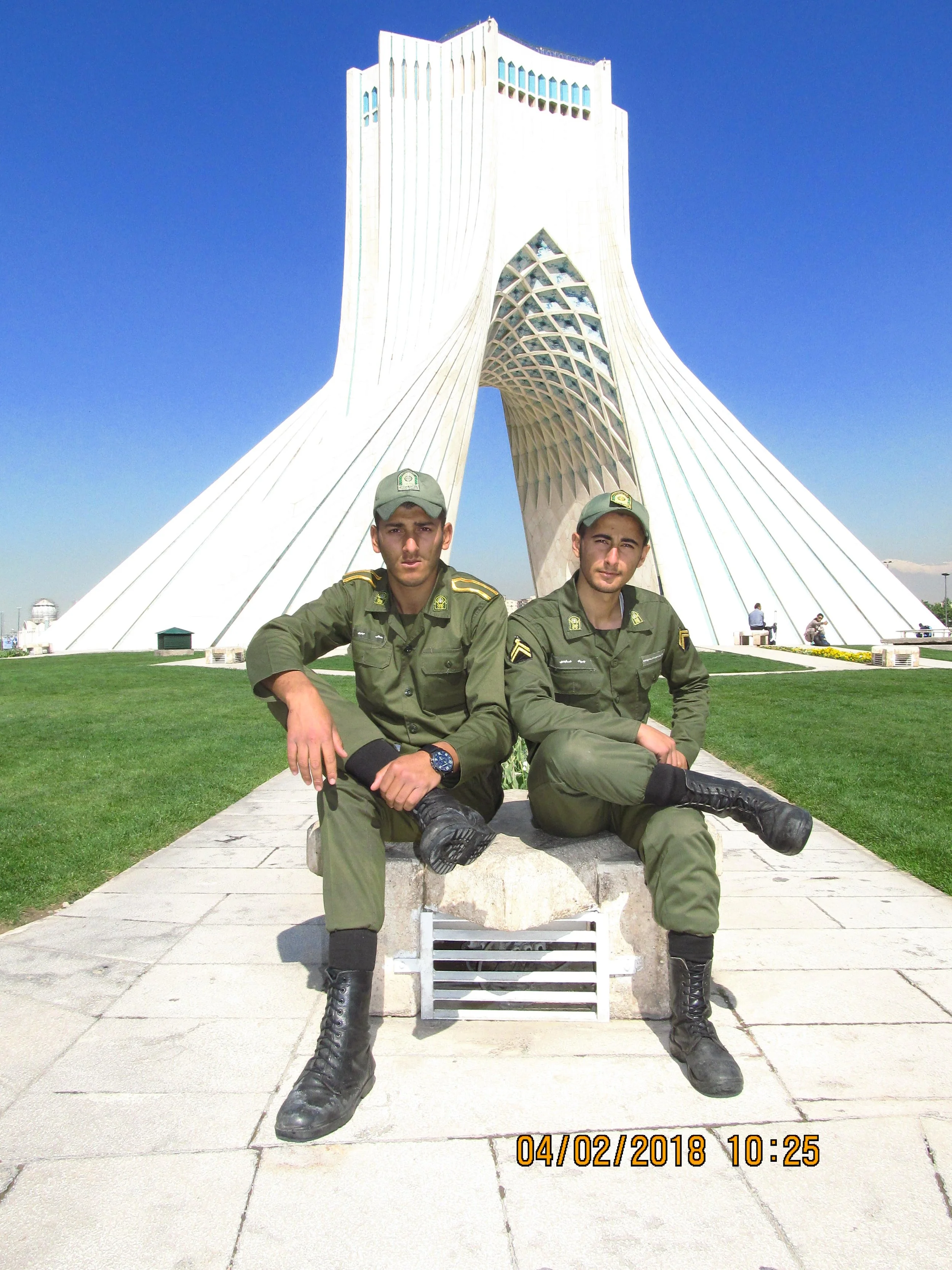 Two uniformed military personnel sitting on a bench in front of the Azadi Tower in Tehran, Iran, with a clear blue sky overhead.