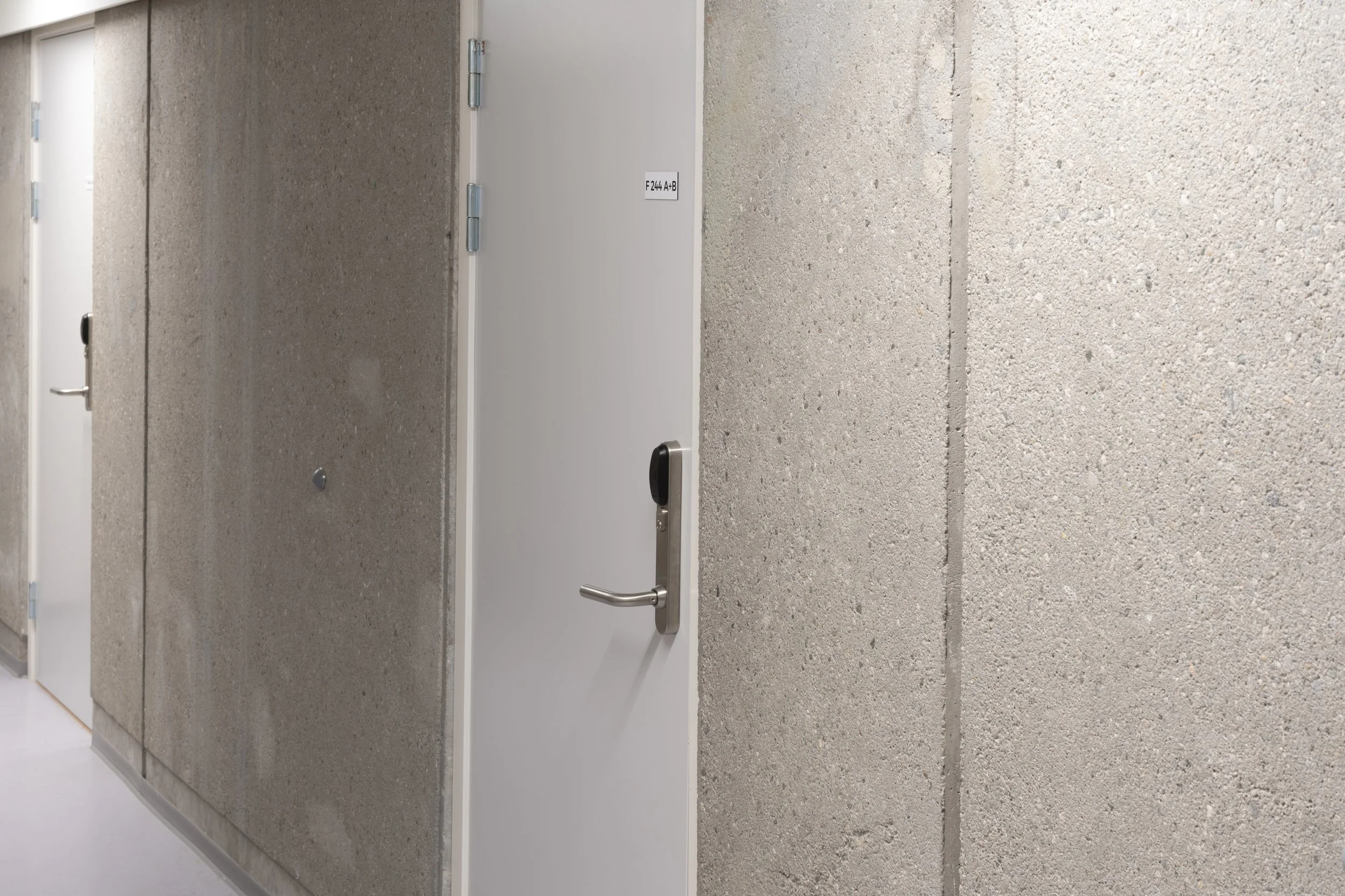 Hallway with concrete walls and white doors, one door with a keypad lock and a small sign that reads 'F244 A+B'.