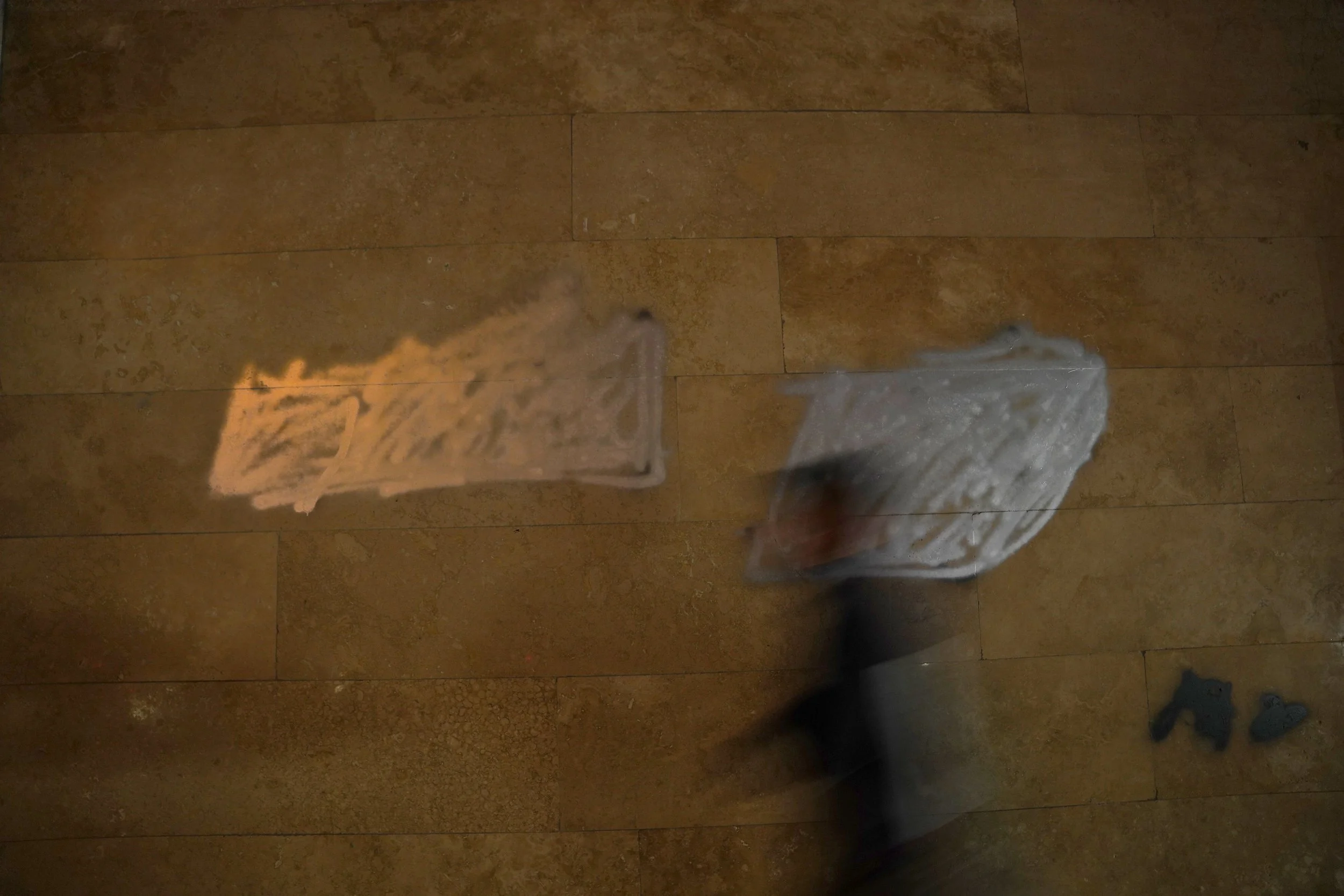 A person is seen cleaning a mirror with a white cloth, with a reflection of the person and the cloth visible in the mirror. The floor is tiled with brownish tiles, and a pair of black shoes are on the floor in the bottom right corner.