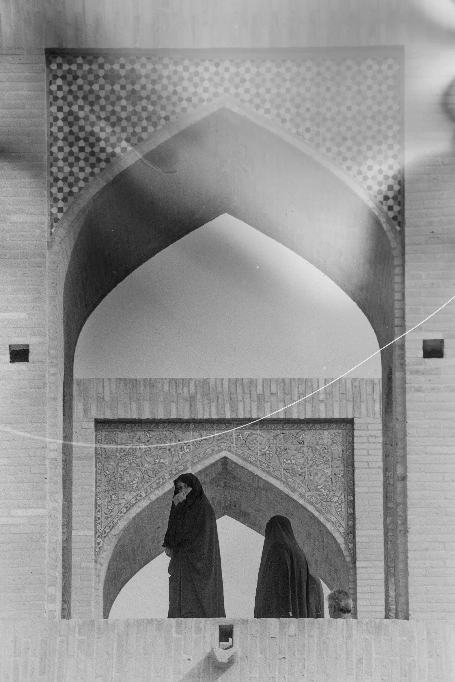 Two women in hijabs standing on a balcony with traditional Islamic architecture, including arches and decorative tile work.