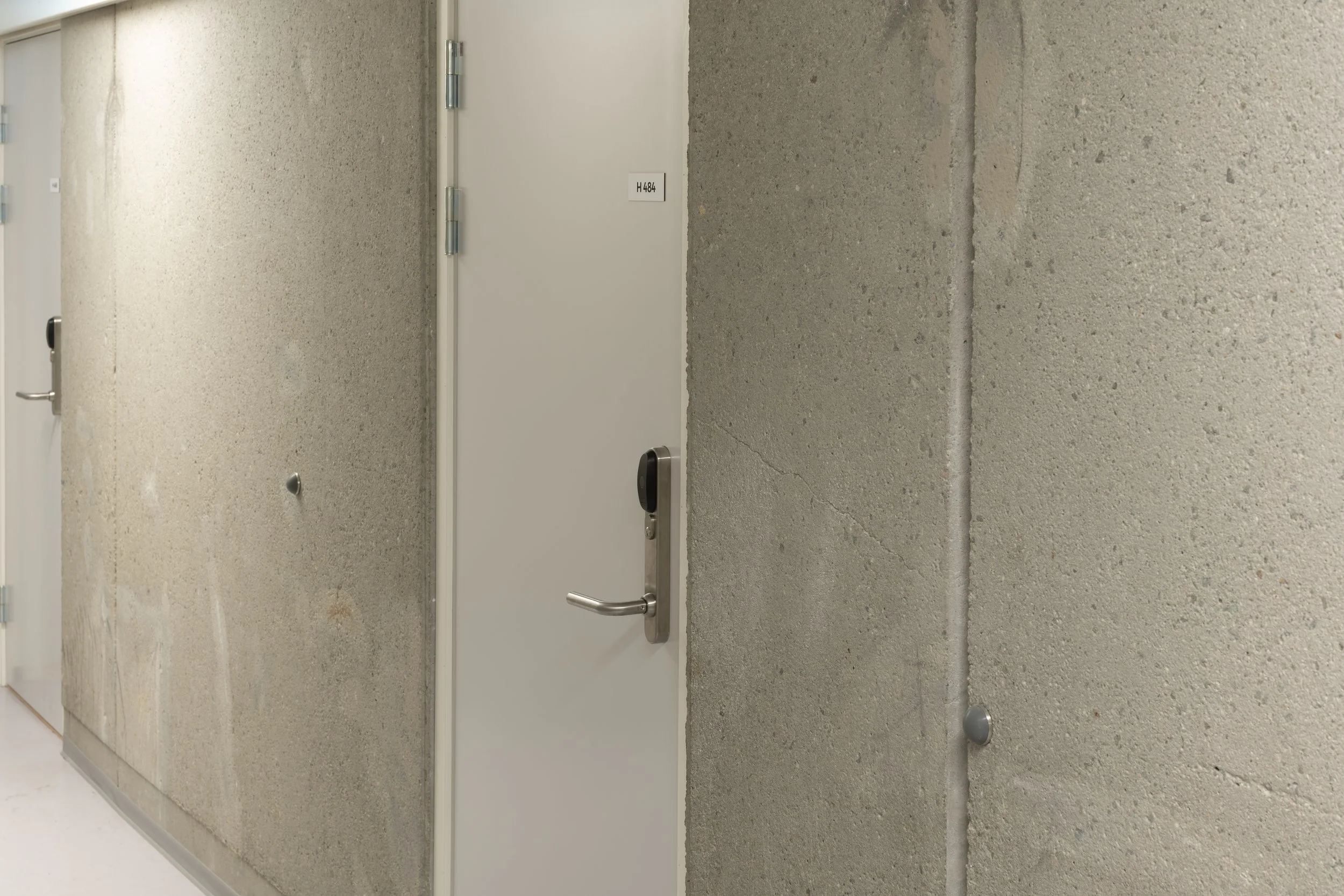 A plain door with a black handle and a small white label, set into a concrete wall, with another door partially visible to the left.