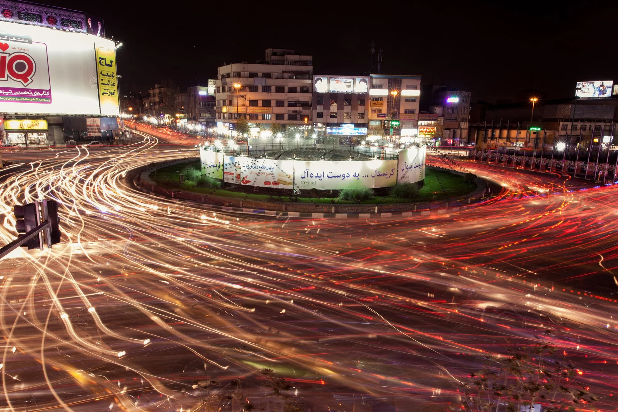 Long exposure photo of a busy city roundabout at night with light trails from moving vehicles and illuminated billboards and buildings surrounding it.