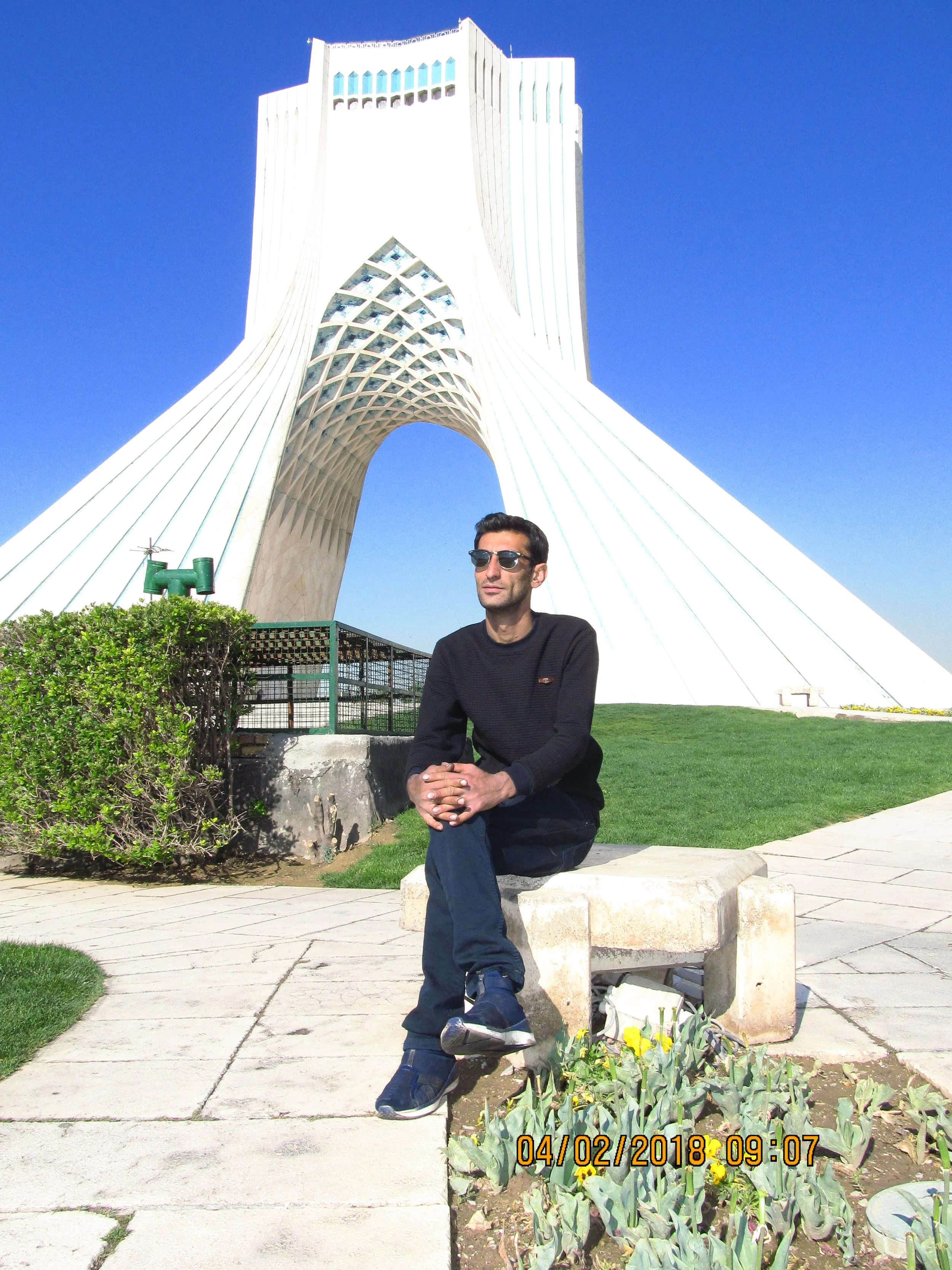 A man sitting on a stone bench in front of Azadi Tower in Tehran, Iran, under a clear blue sky, wearing sunglasses and a black sweater.