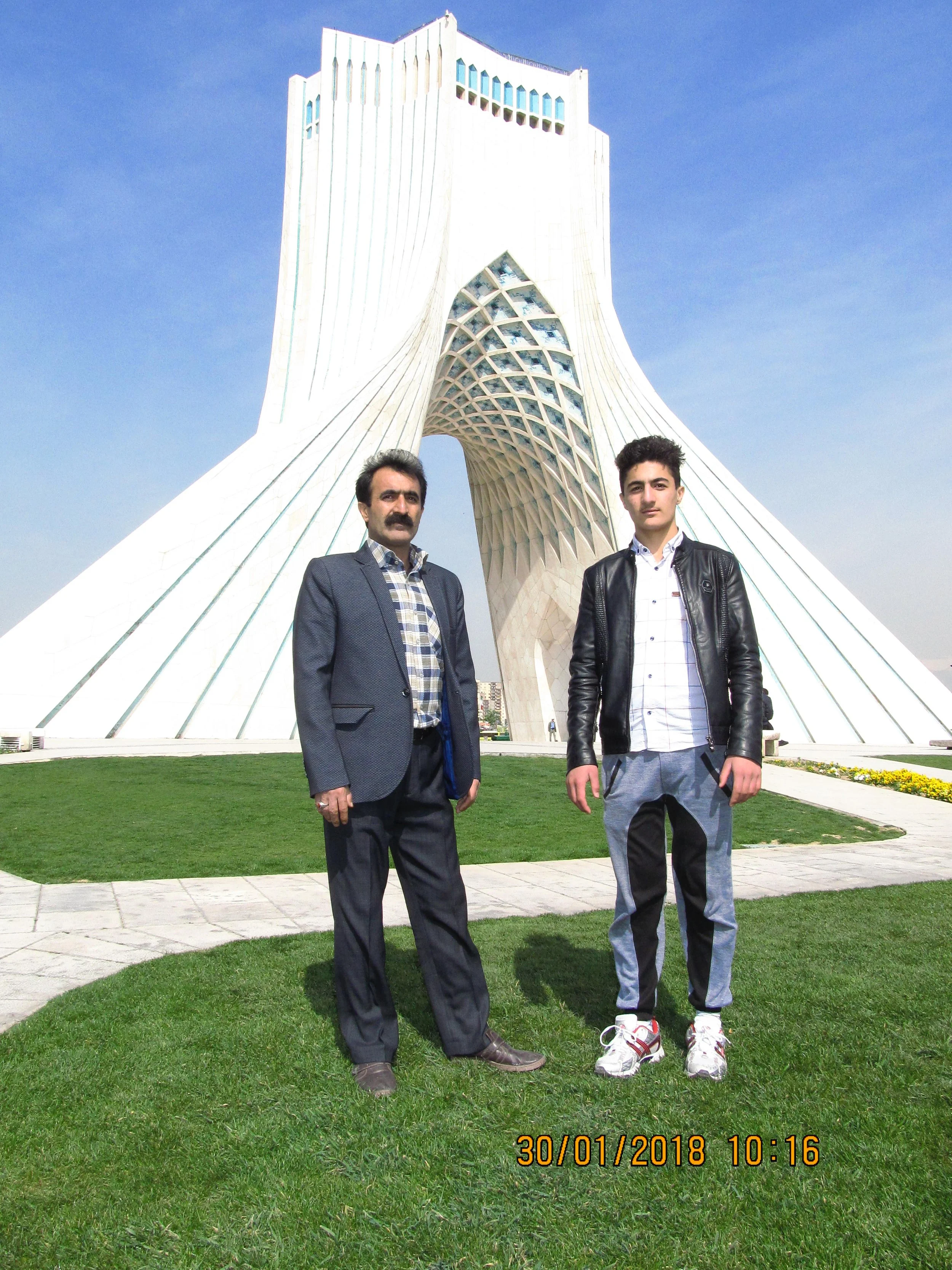 Two men standing on a grassy area in front of a tall, modern white architectural building with an arched opening and unique design, under a clear blue sky.