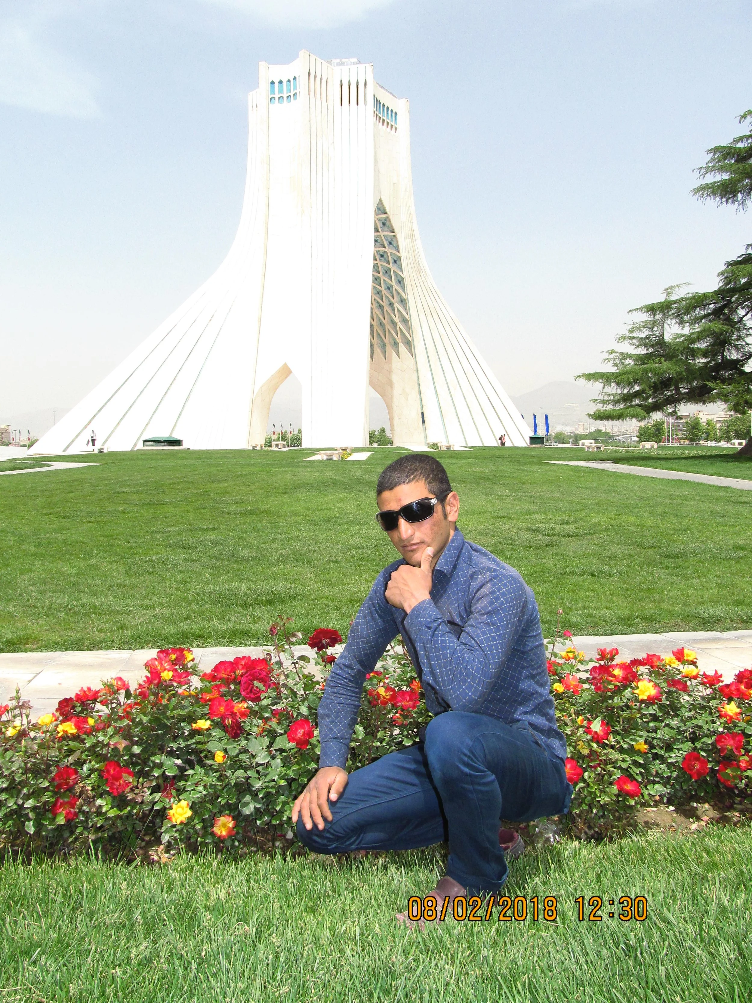 A man in sunglasses crouching near a flower bed of red and yellow flowers in front of a large white building with curved architecture, on a sunny day.