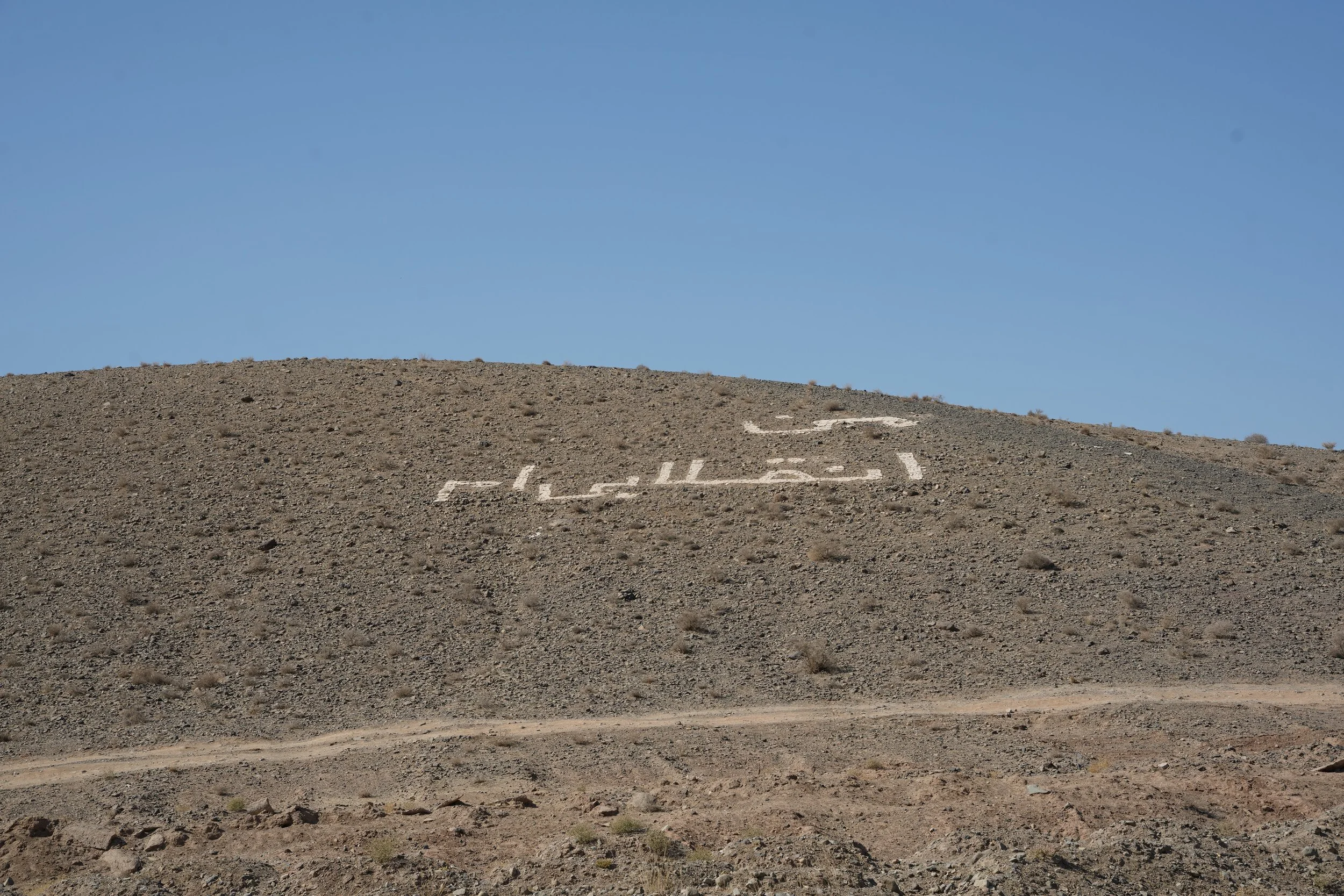 Desert hill with white Arabic writing on it under a clear blue sky.