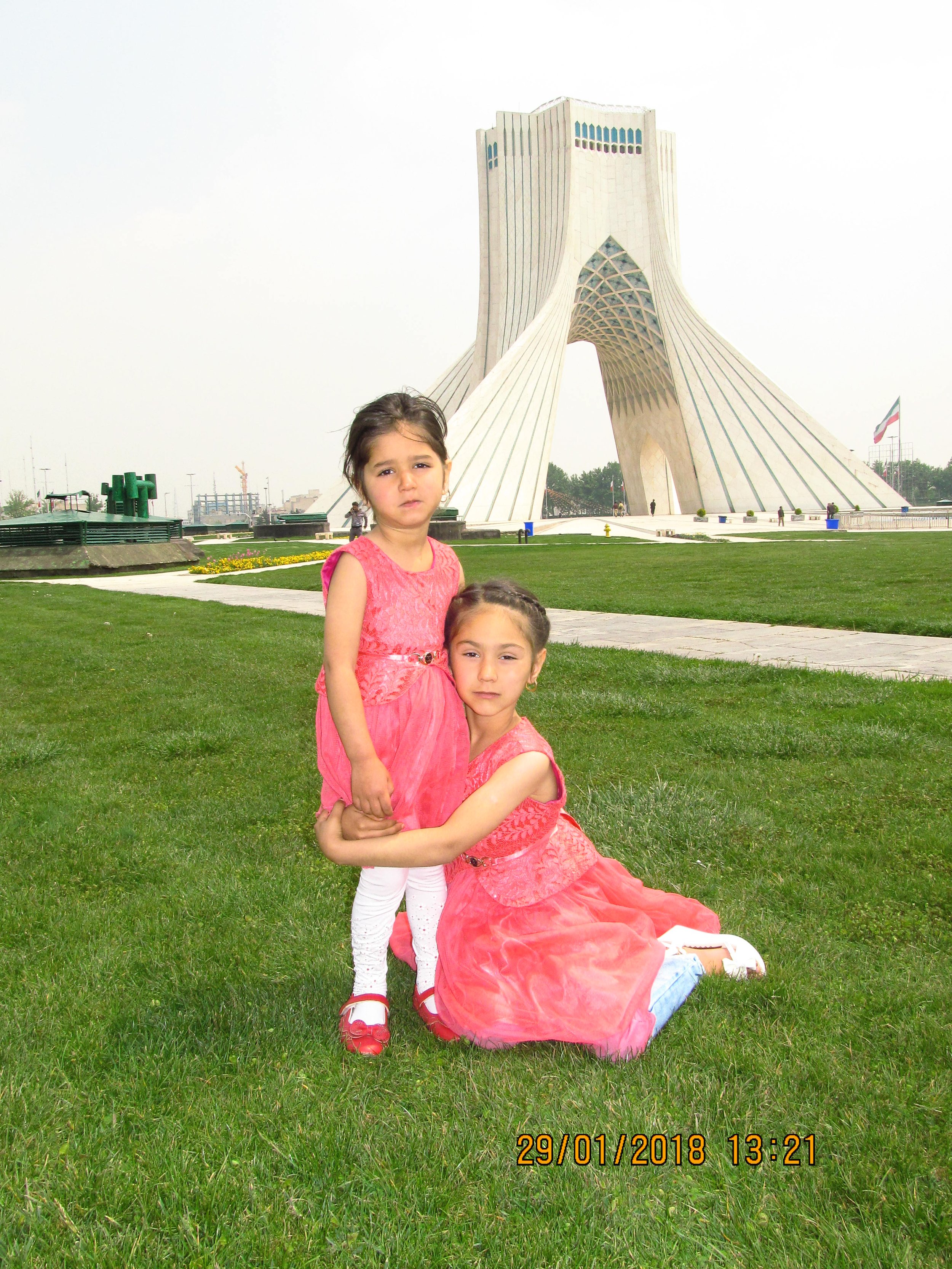 Two young girls in pink dresses are outside on grass in front of the Azadi Tower in Tehran, Iran.