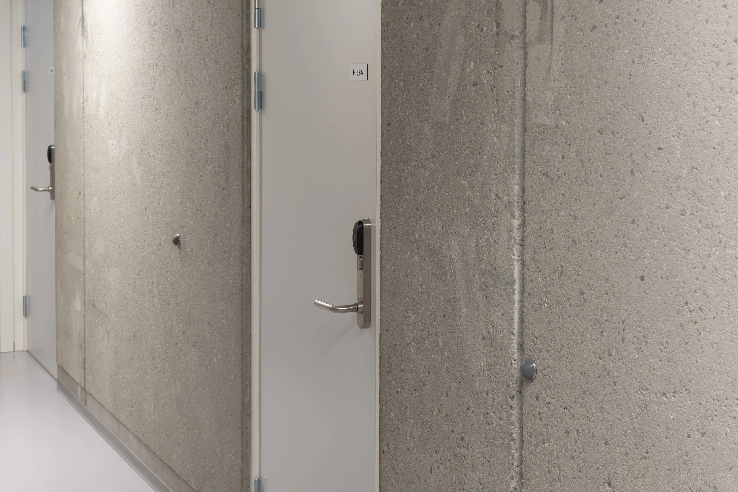 A hallway with concrete walls and a closed gray door with a silver handle and electronic lock.