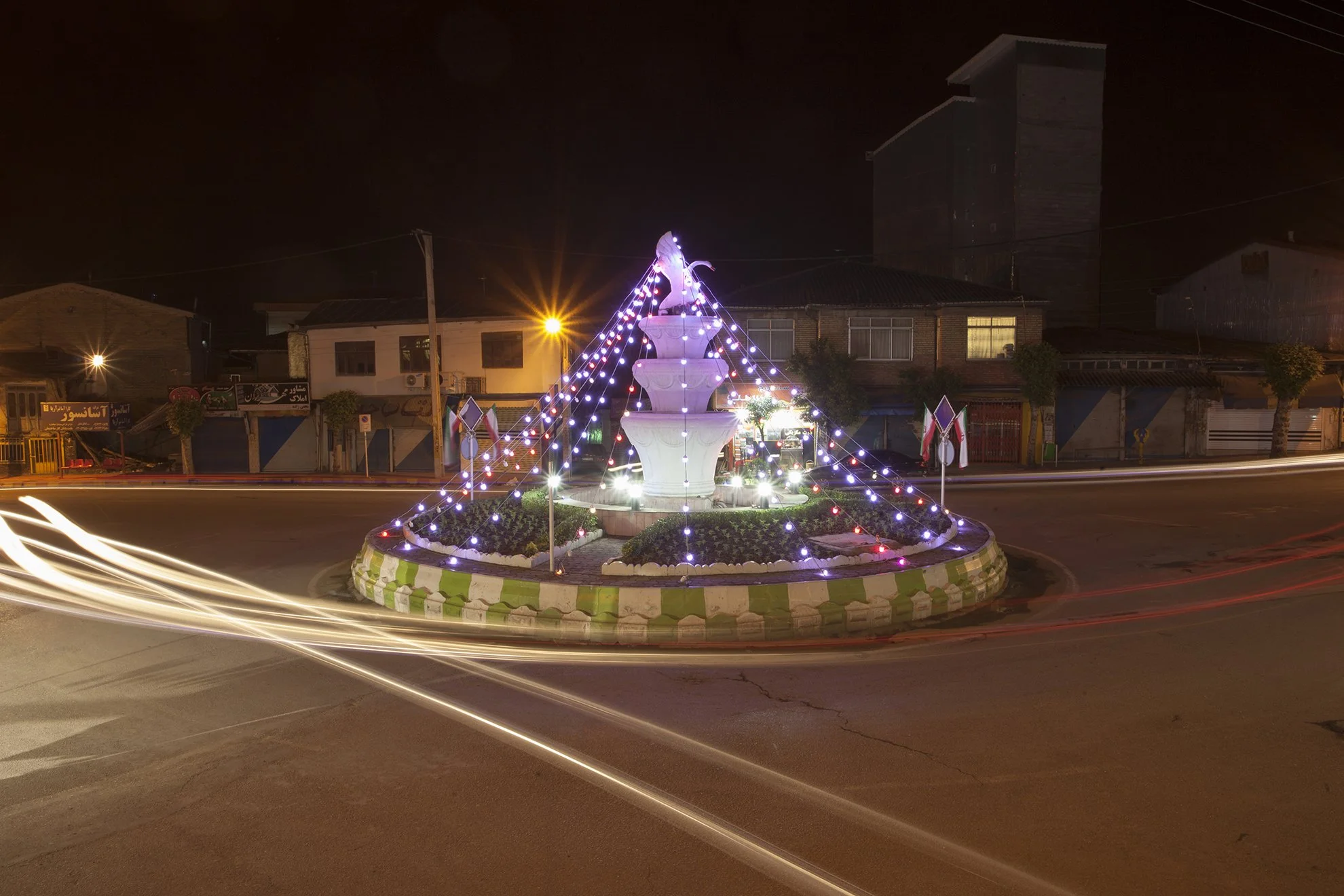 Night scene of a decorated roadside roundabout with a white fountain in the center, wrapped in colorful string lights and small flags, with blurred light streaks from passing vehicles.