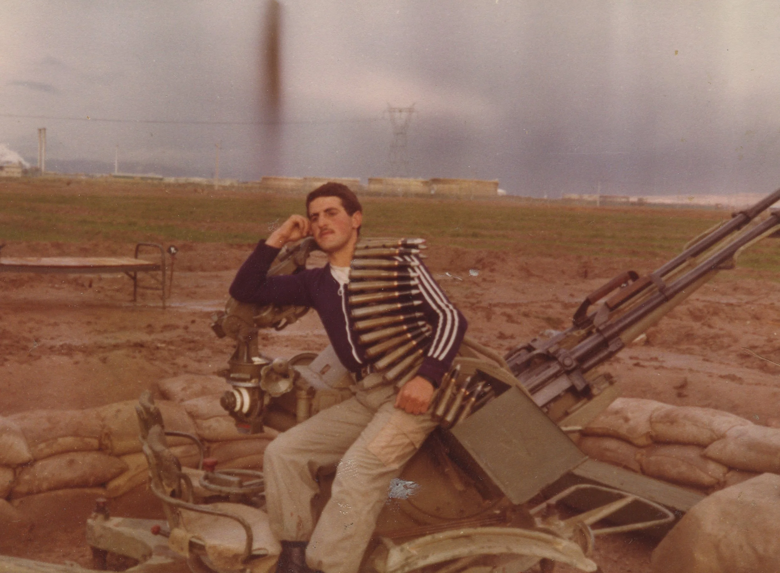 A man with dark hair, wearing a dark sweater with white stripes, sitting on a military vehicle, resting his head on his hand with a serious expression. The vehicle has a mounted weapon with ammunition belts, sandbags, and a cloudy sky in the background.