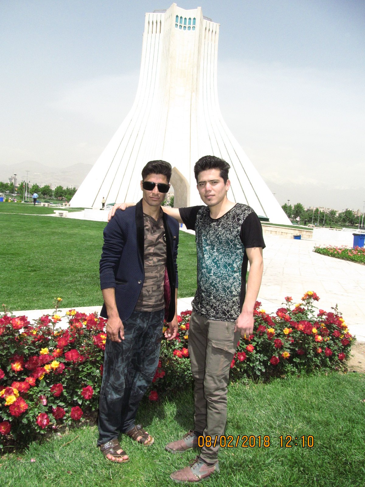 Two men pose in front of a large white monument with flower beds and green grass around them, under a partly cloudy sky.