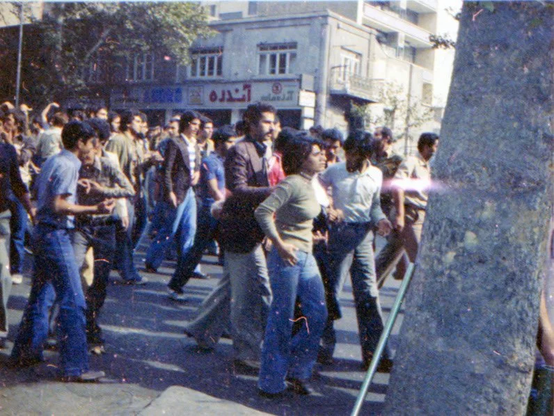 Group of people walking across a city street, with storefronts in the background and a large stone structure on the right.