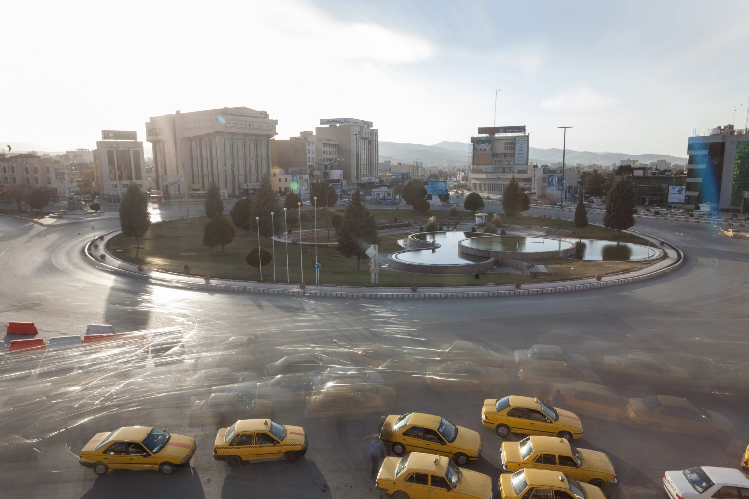 A city square with a roundabout, landscaped with trees and a water feature, surrounded by tall buildings and a parking lot filled with yellow taxis and cars, during daytime with sunlight.