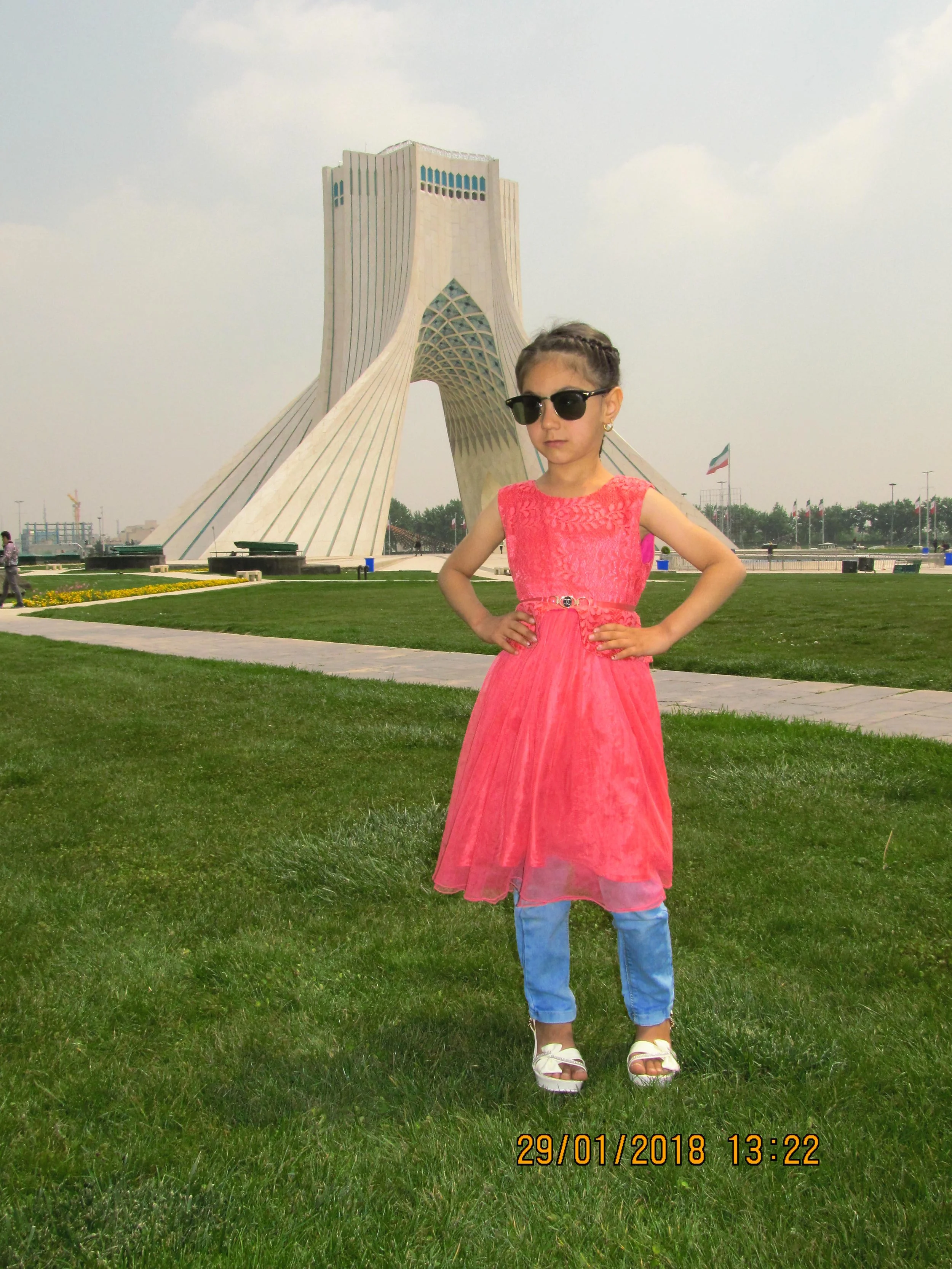 A young girl in a pink dress and blue jeans standing on grass with hands on hips, wearing sunglasses, and a modern architectural building in the background.