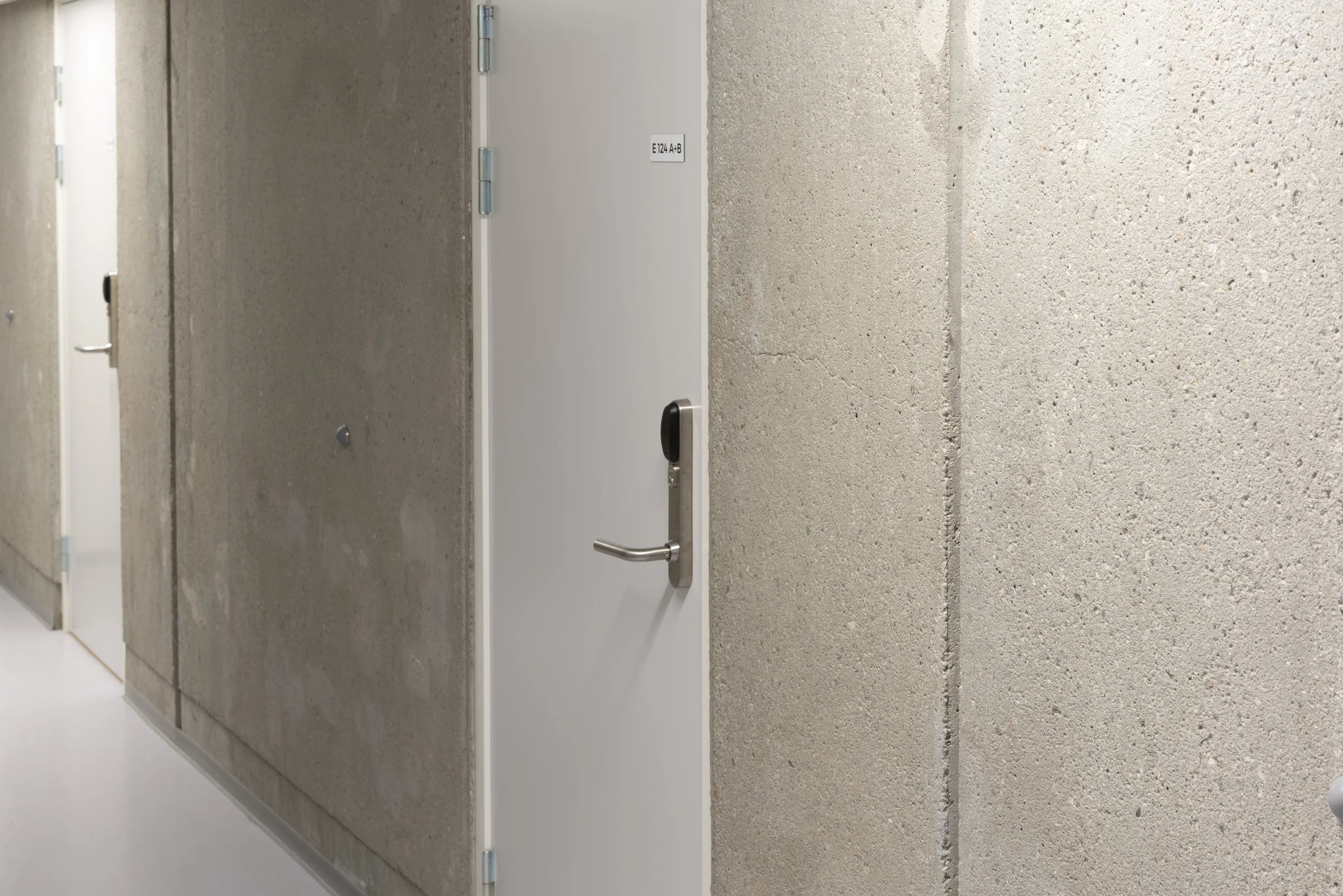 A hallway with concrete walls and white doors, one of which has a black handle and a small sign labeled 'E124 A-B'.
