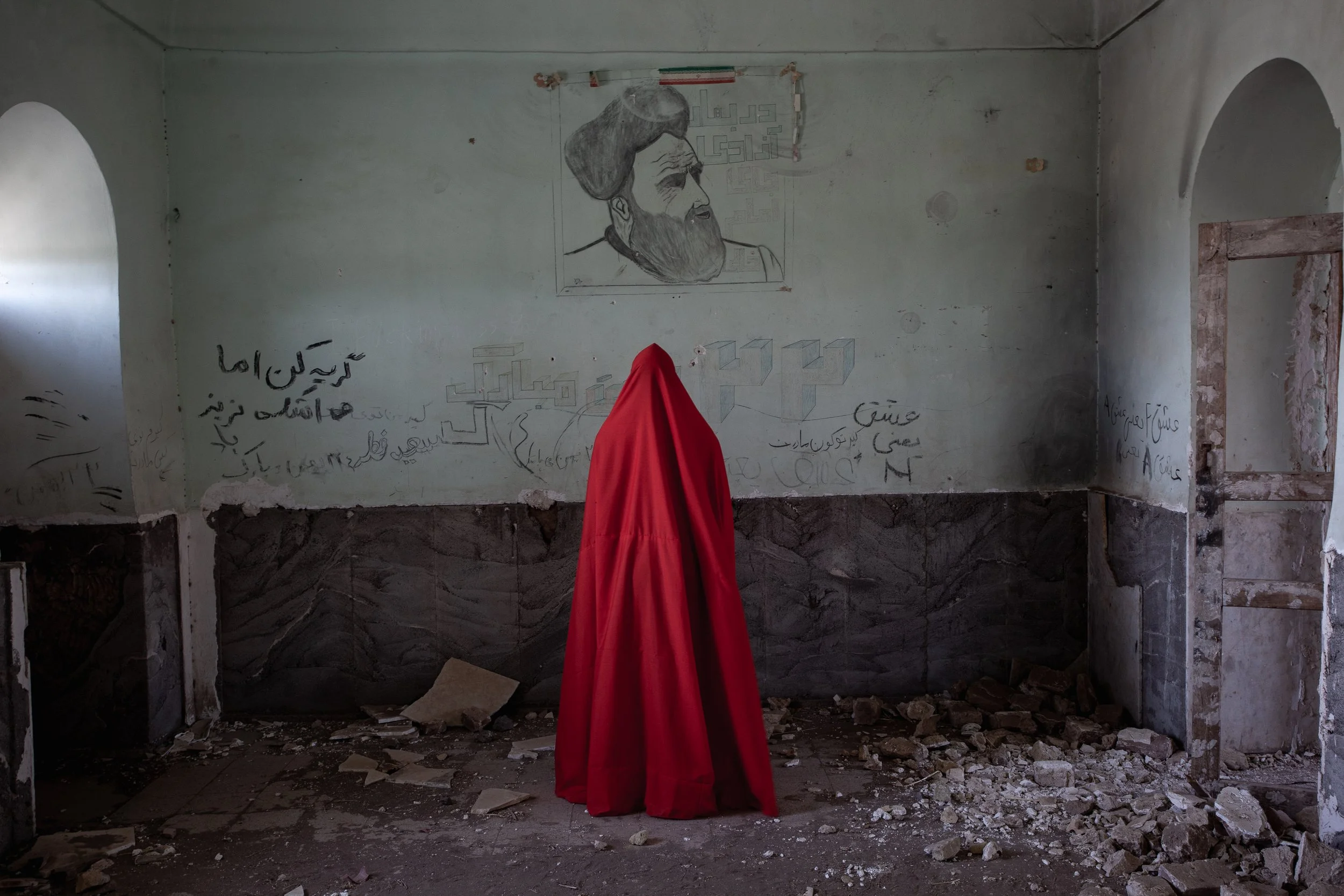 A person draped in a red cloak stands inside an abandoned, dilapidated room with bare concrete floors and walls. The room has graffiti, sketches, and a portrait of a man on the wall.