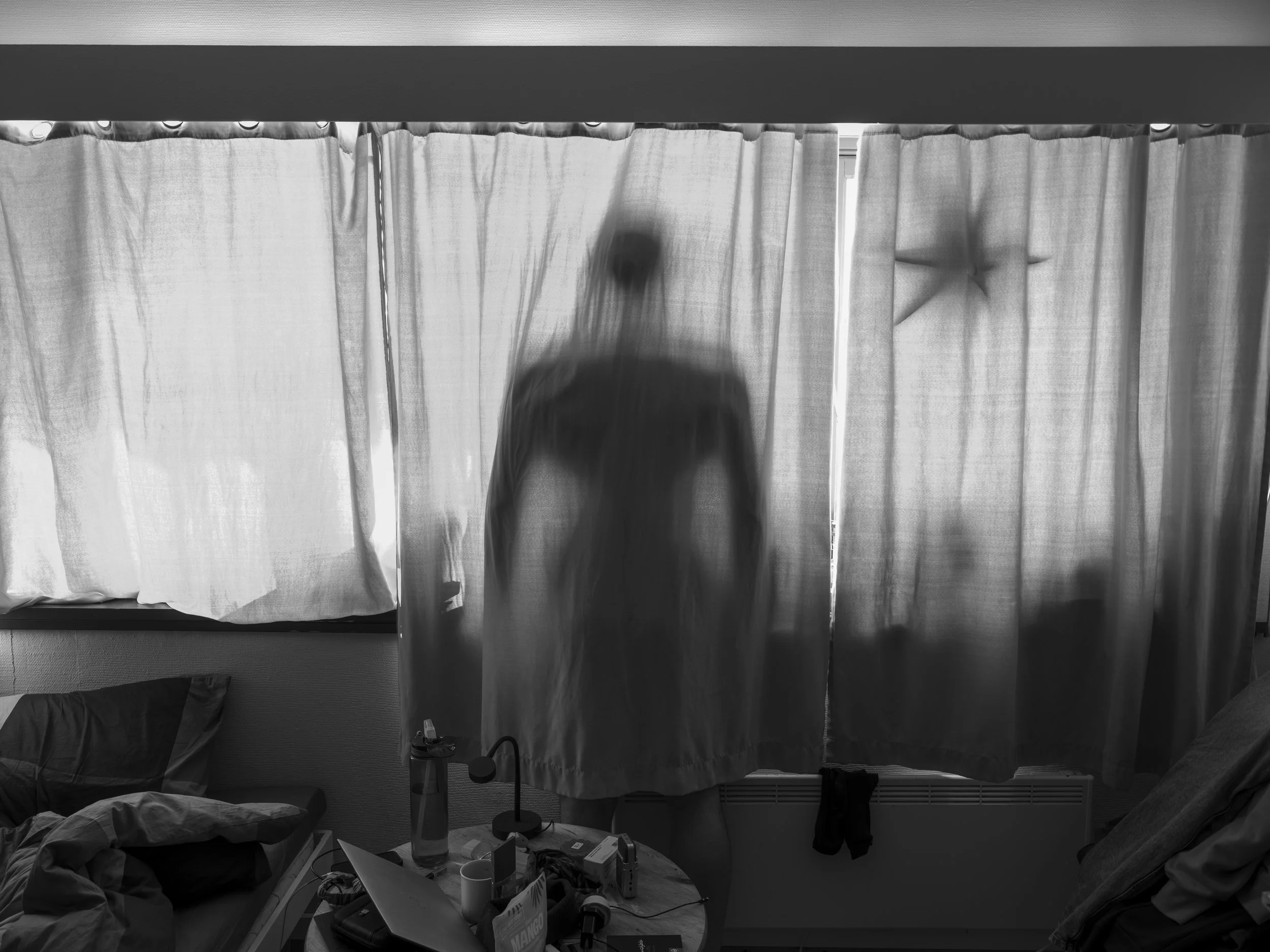 A silhouette of a person behind a semi-transparent curtain in a dimly lit room, with a bed, cluttered table, and window with curtains in the background.