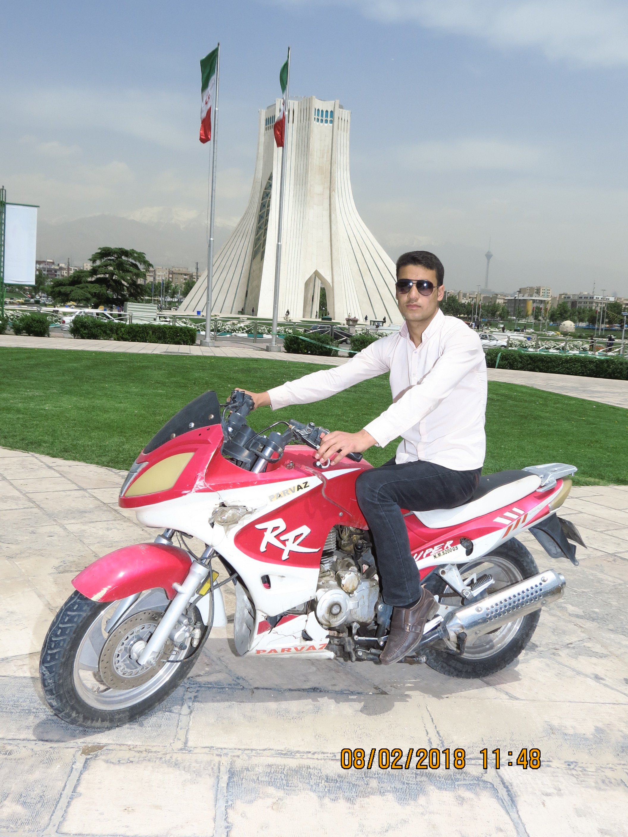A man wearing sunglasses and a white shirt sitting on a red and white motorcycle in front of the Azadi Tower in Tehran, Iran, with flags in the background and a timestamp of February 8, 2018, at 11:48.