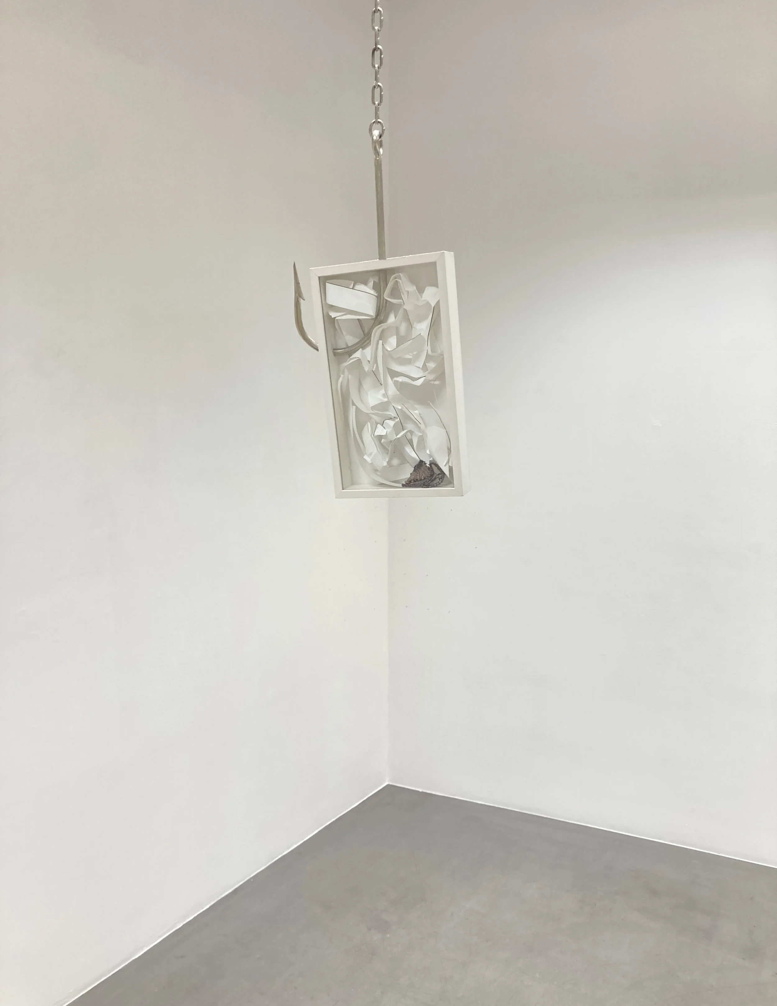 White art piece with crumpled paper inside a rectangular frame hanging from a chain on a white wall corner.