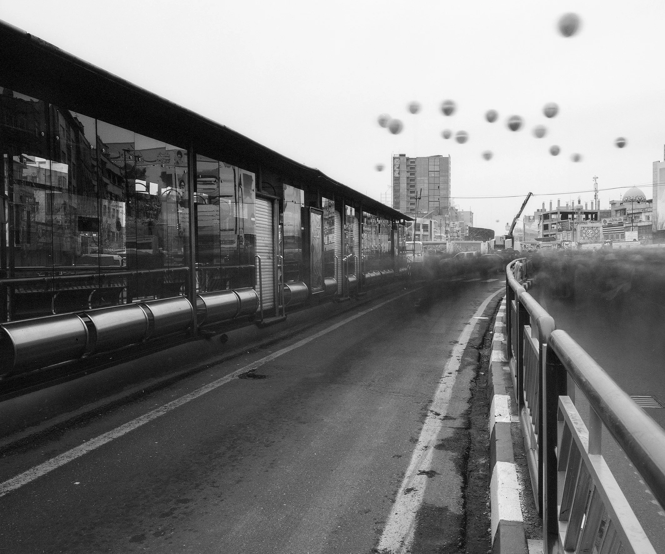 Black and white photograph of a city street with a bus stop, glass bus shelter, and city buildings in the background.