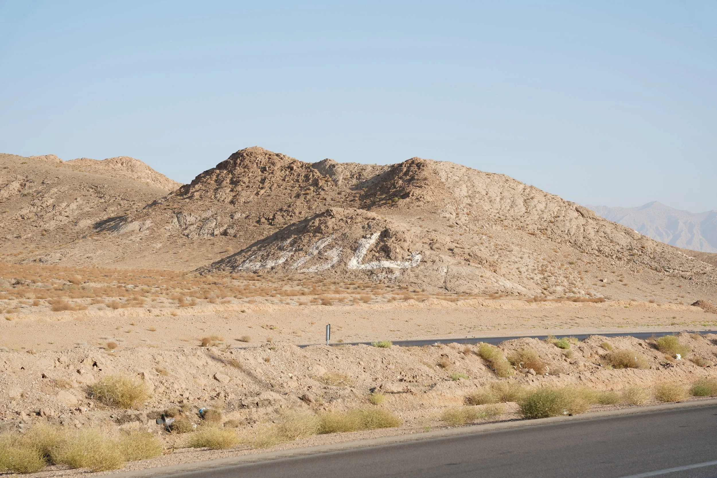 Desert landscape with a rocky hill that has the word 'LOVE' painted in white on it. Dry bushes and a road are in the foreground.