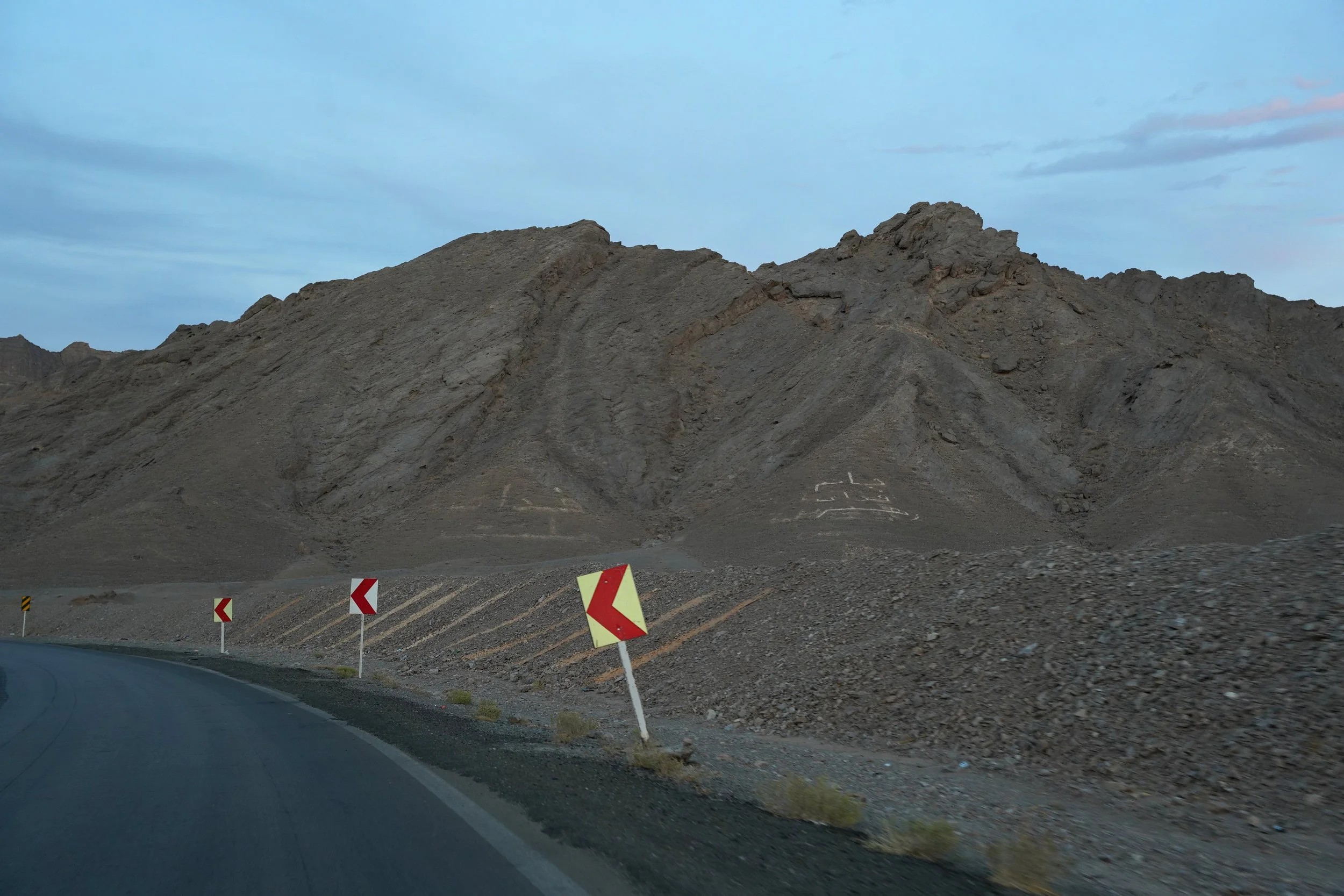 A winding mountain road with red and white directional signs on the side, passing through a barren, rocky landscape with mountains and an overcast sky in the background.