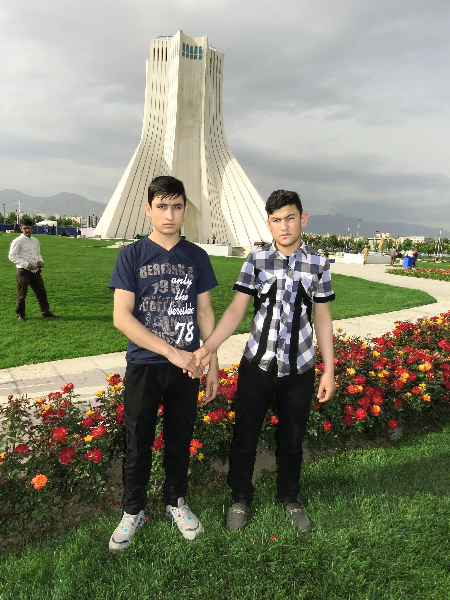 Two young boys standing together in front of a large monument, holding hands, on a grassy area with flowers and a cloudy sky.