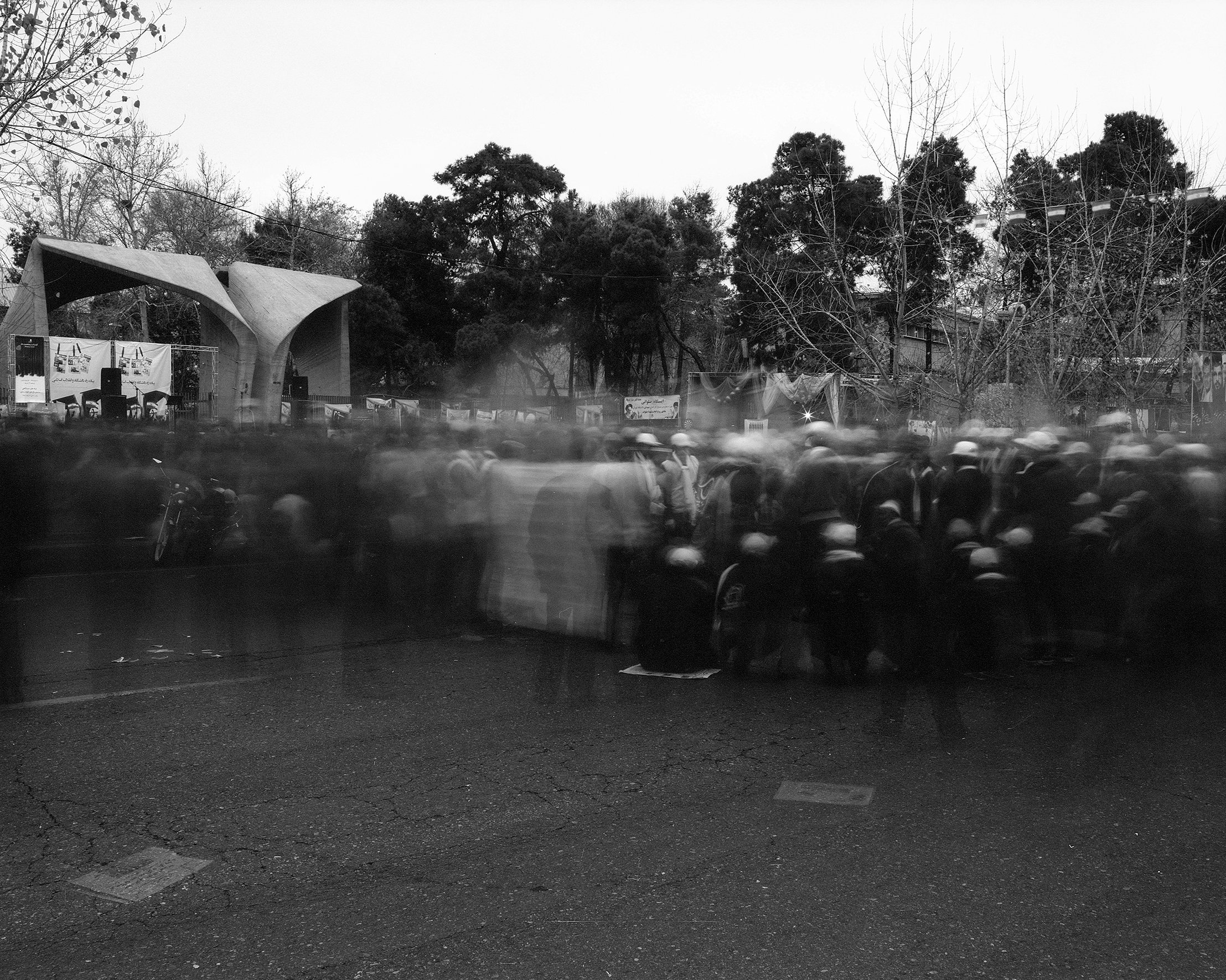Black and white photo of a large group of people standing outdoors, with some wearing helmets. In the background, there is a stage with musical instruments, banners, and trees.