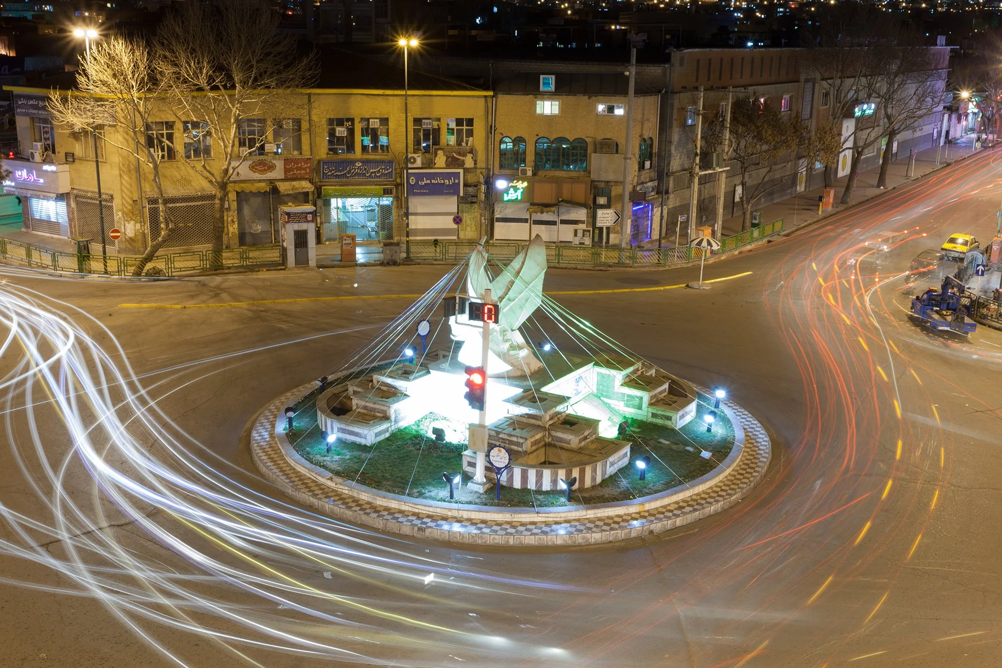 Nighttime view of a city roundabout with a lit-up central monument and light trails from moving vehicles, surrounded by buildings with illuminated signs and leafless trees.